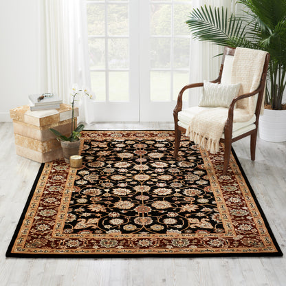 Black oriental area rug with gold and red border placed in a bright living room beside a wooden chair and potted plants.