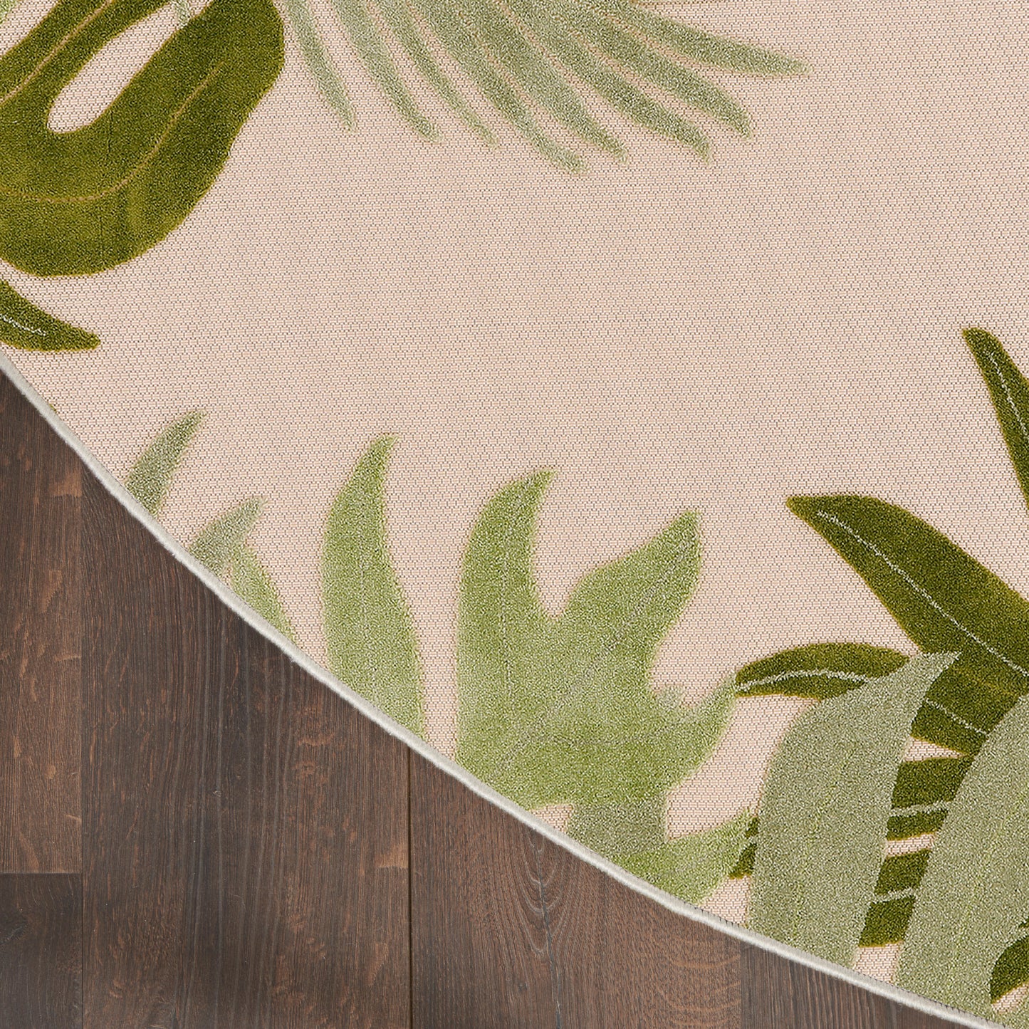 Round beige rug with green tropical leaf print and white edging on dark wooden floor.