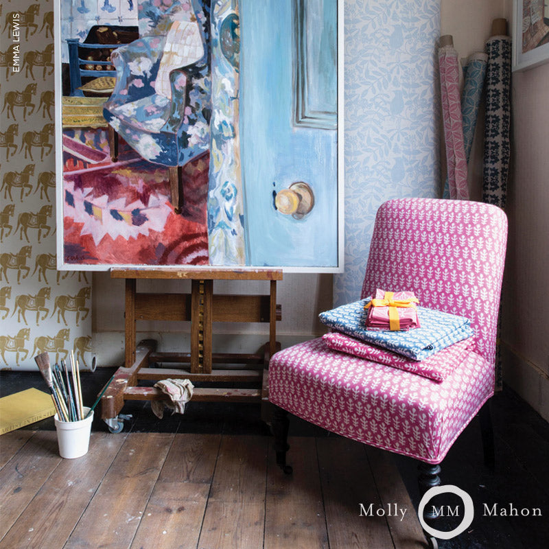 Pink patterned upholstered chair beside an easel with a colorful painting, stacked fabrics and wrapped gift on chair.