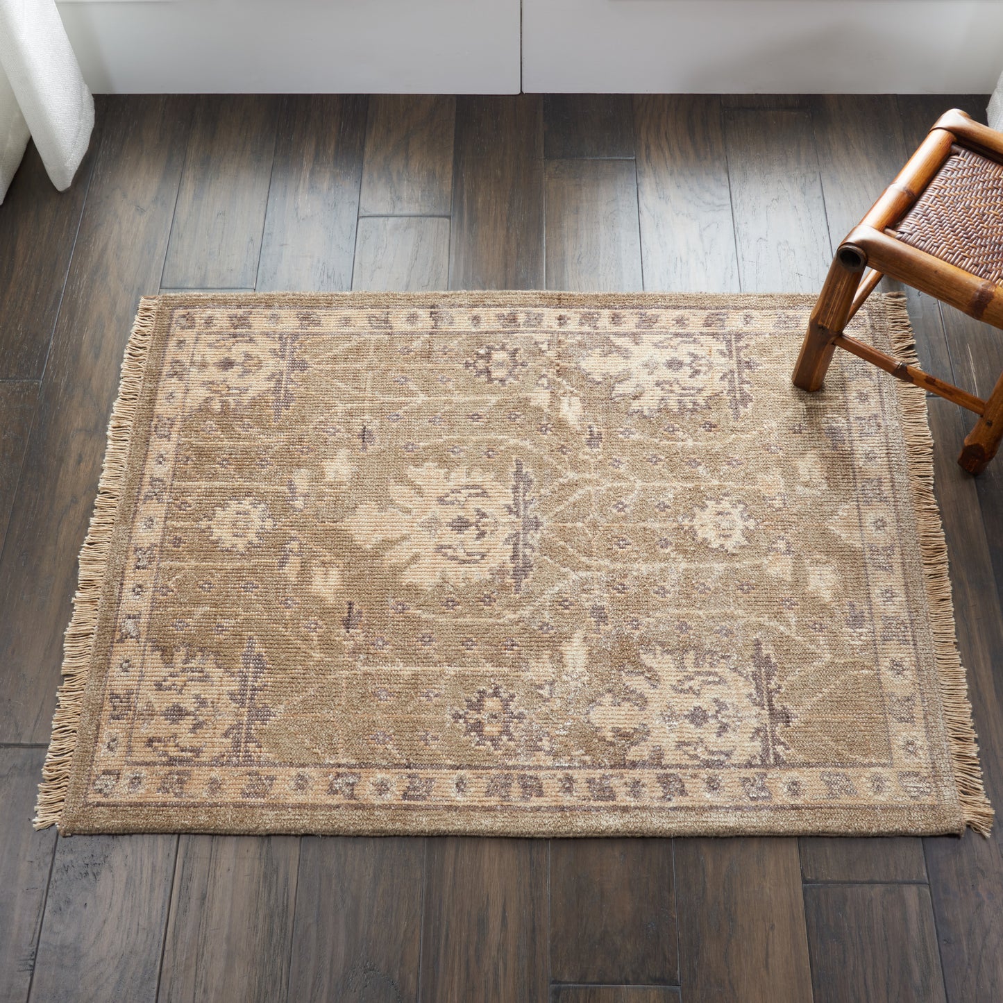 Beige oriental-style rug with floral motifs and fringed borders on dark hardwood floor, near a wicker chair.