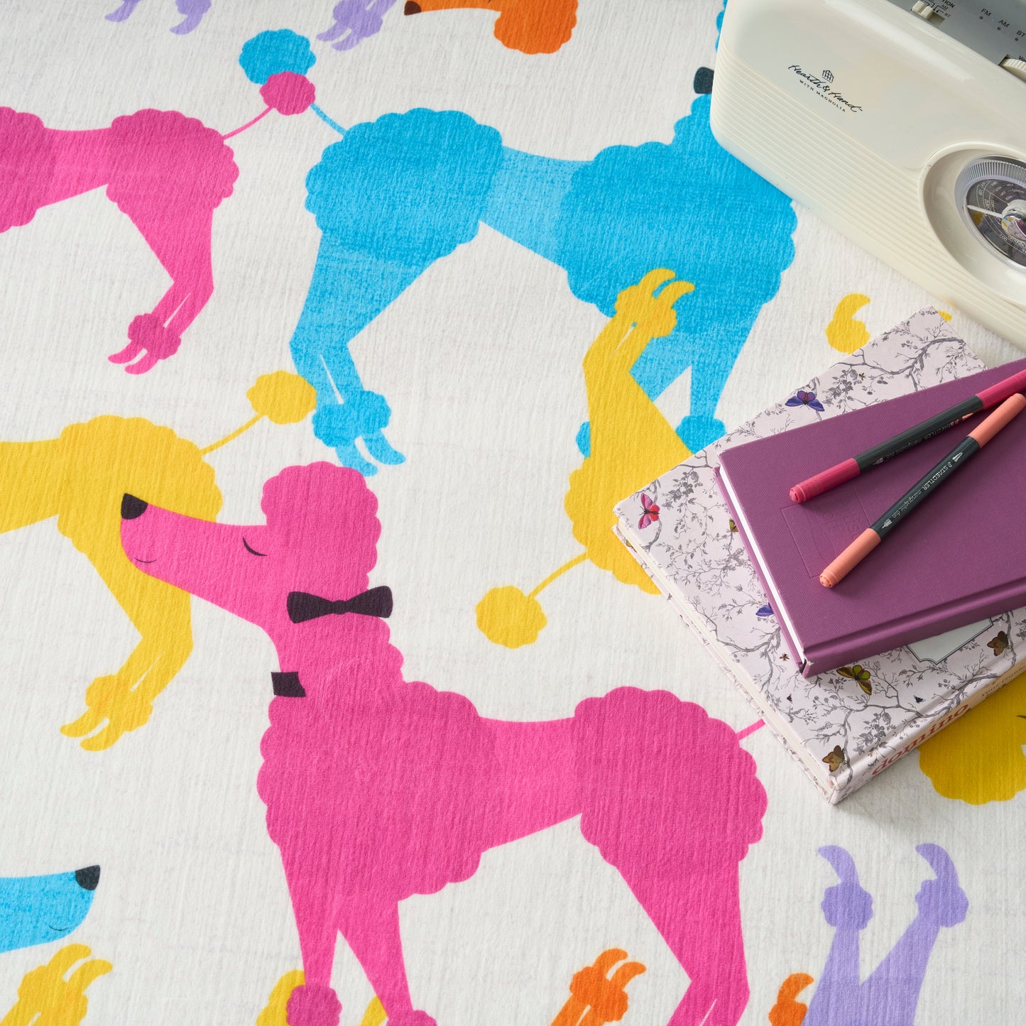 Bright poodle silhouette pattern in pink, blue, yellow on fabric, with purple notebook, pastel pencils, and a retro white camera.