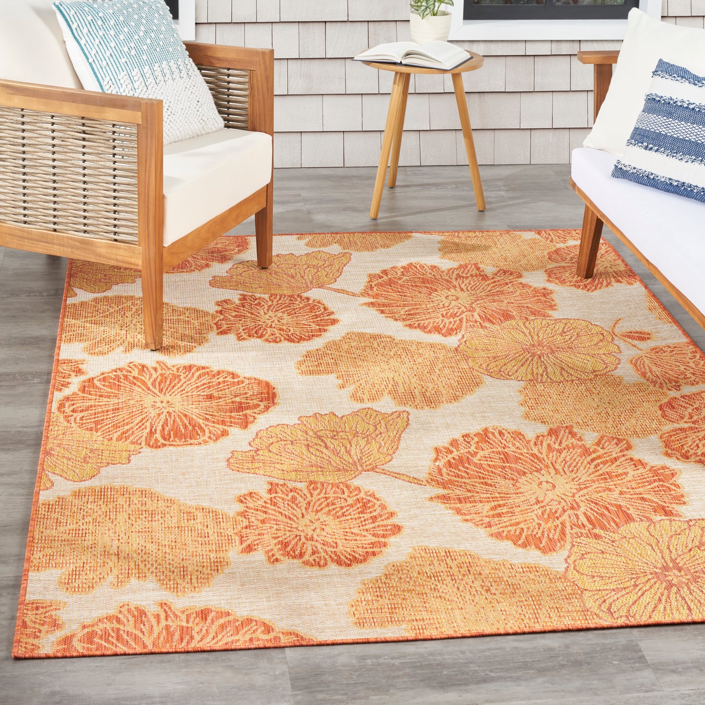 Outdoor seating area with orange floral rug, wooden-frame chairs, white cushions, blue pillows, round side table and book.