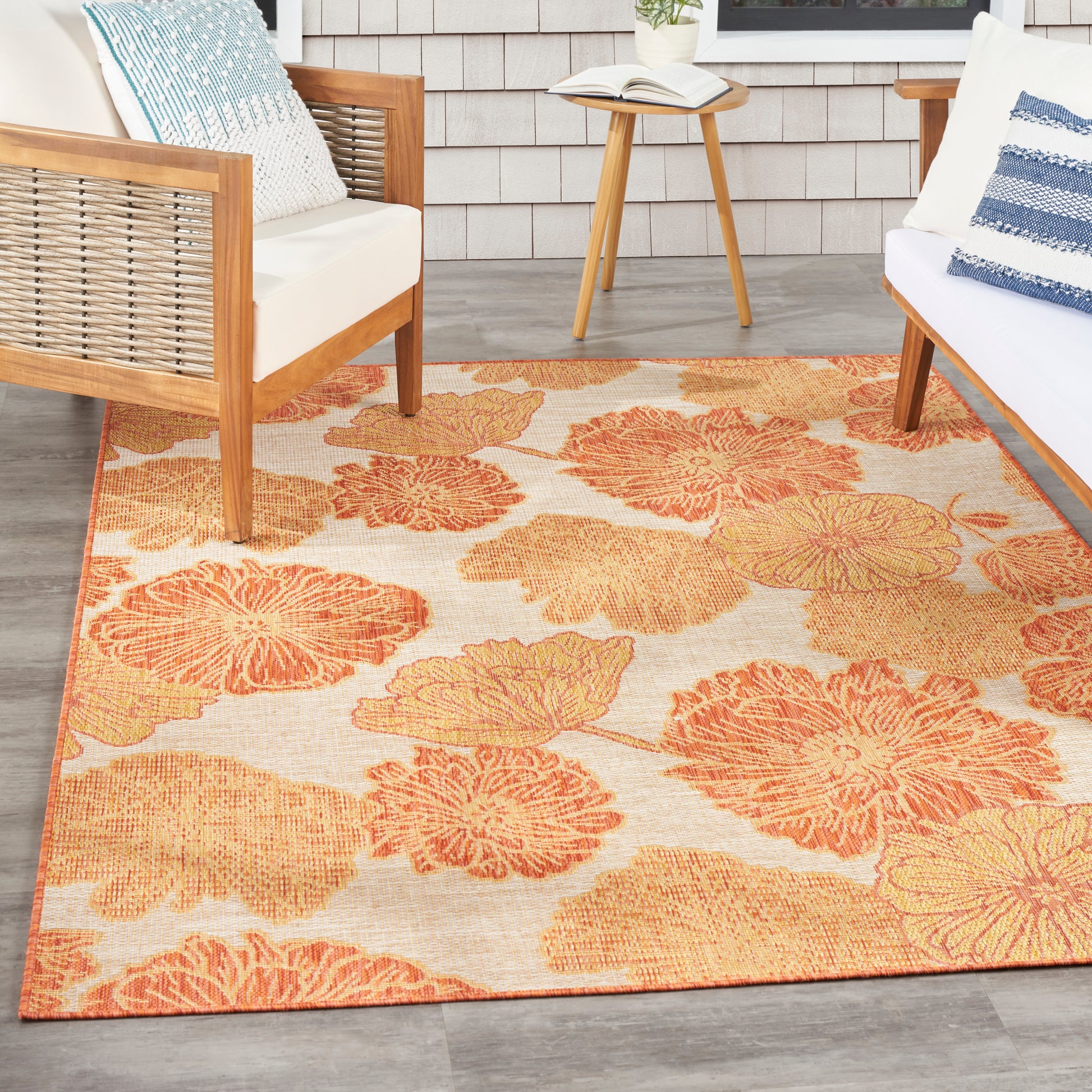 Outdoor seating area with orange floral rug, wooden-frame chairs, white cushions, blue pillows, round side table and book.