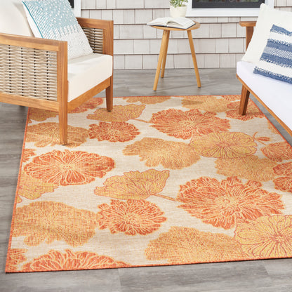 Outdoor seating area with orange floral rug, wooden-frame chairs, white cushions, blue pillows, round side table and book.