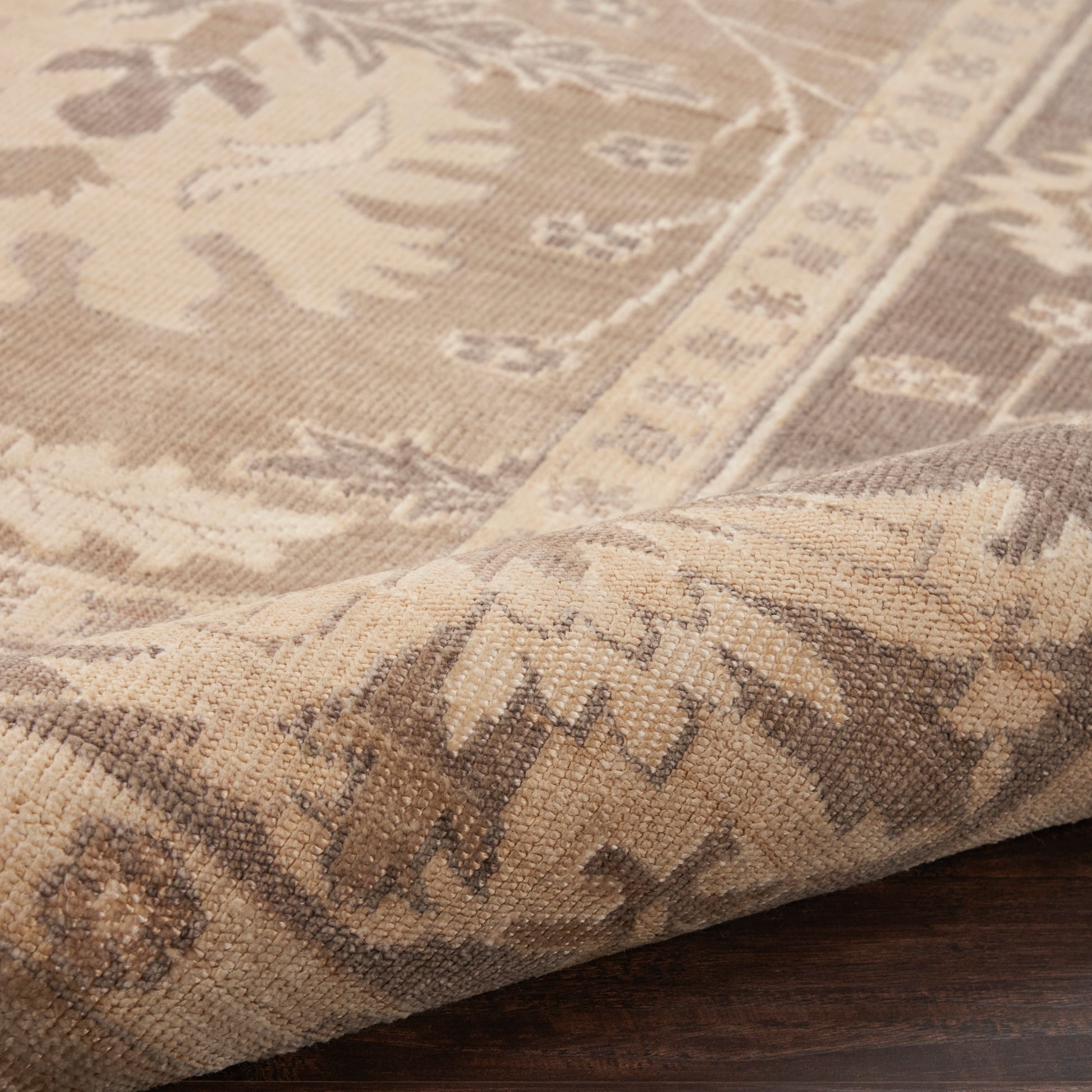 Close-up of a beige and taupe oriental-style rug with a floral border, folded at the corner on a dark wood floor.