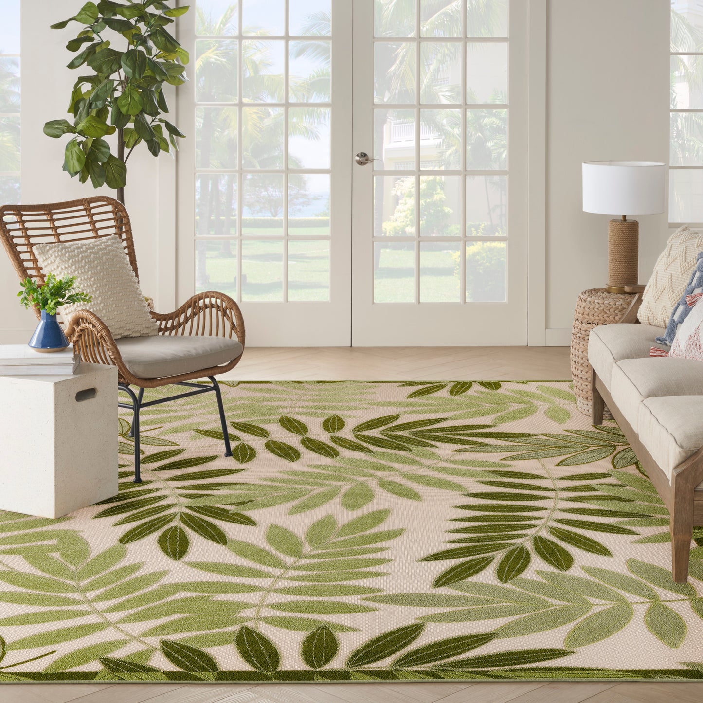 Living room with a green palm-leaf rug, wicker chair, beige sofa, potted plant, and French doors.