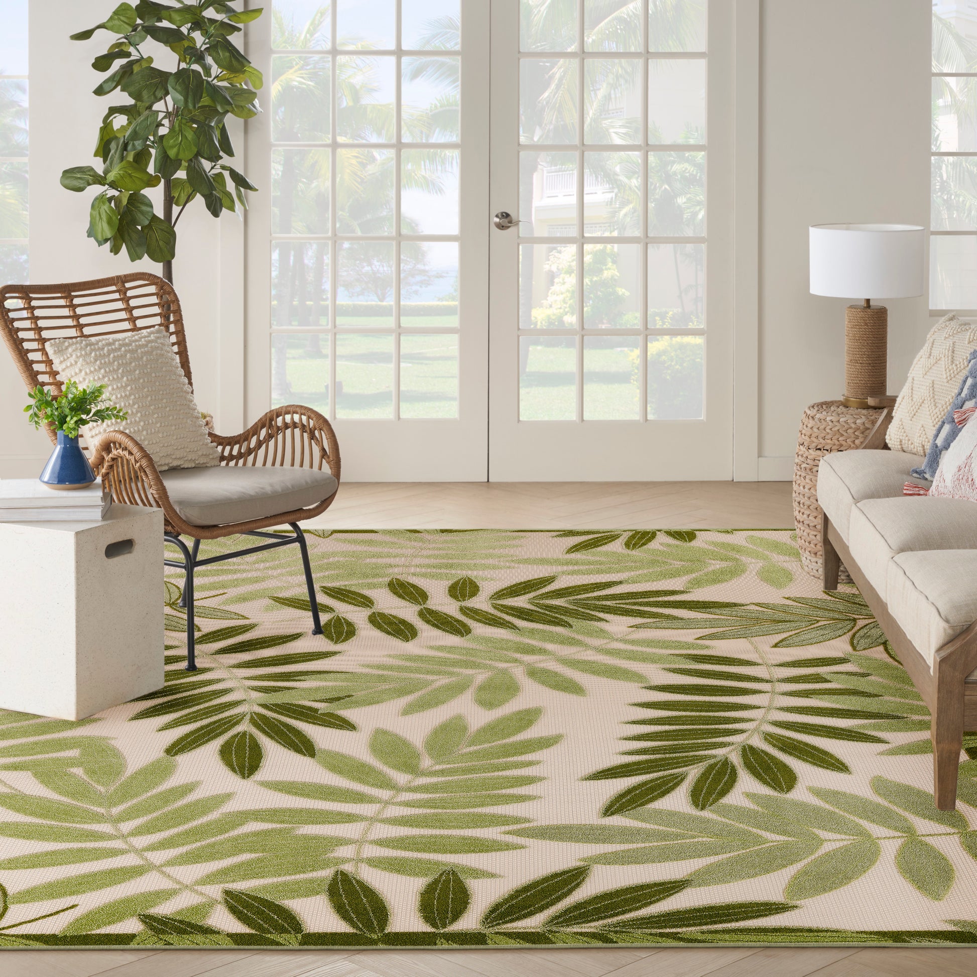 Living room with a green palm-leaf rug, wicker chair, beige sofa, potted plant, and French doors.