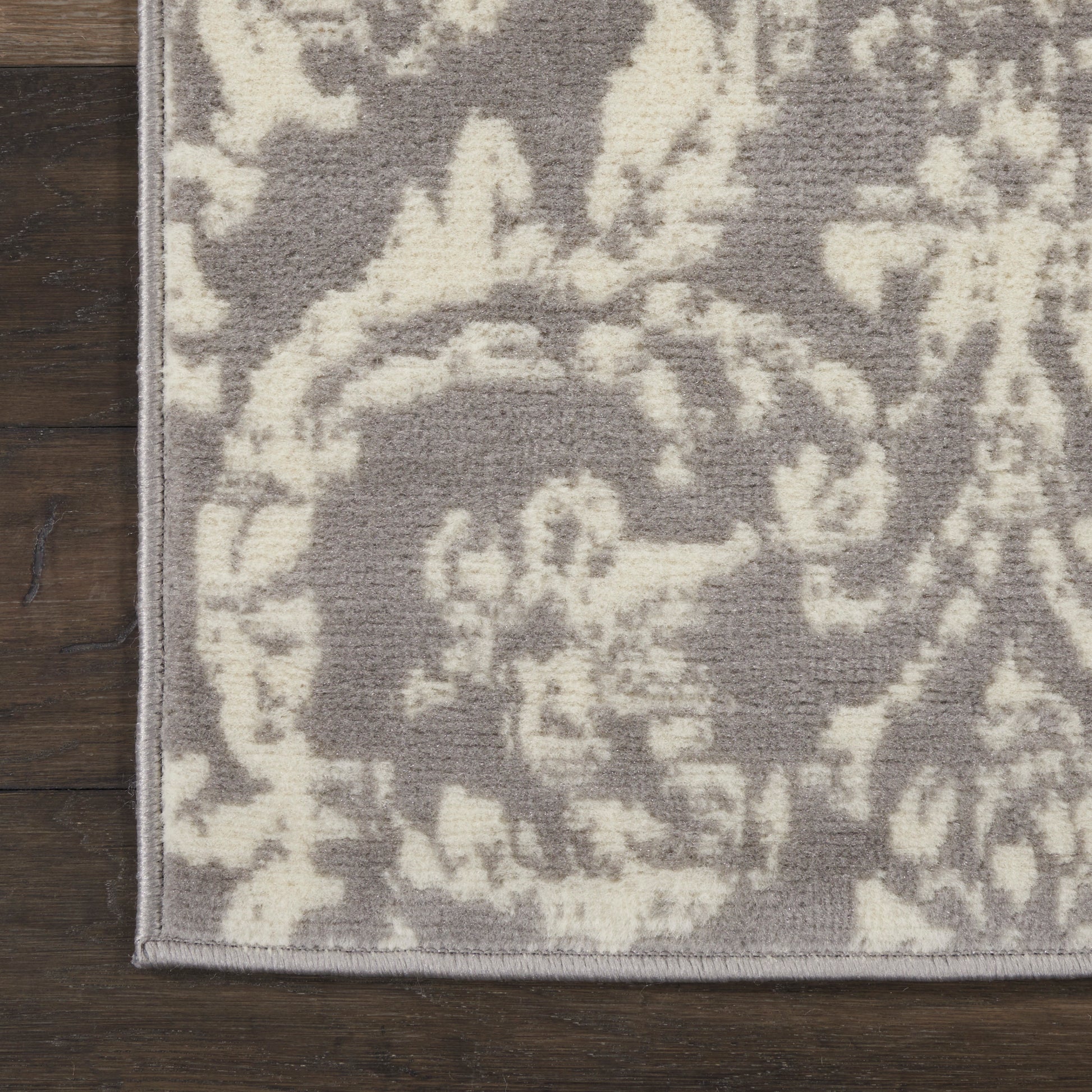 Gray rug with cream damask floral pattern, bound edge, on dark wood floor.
