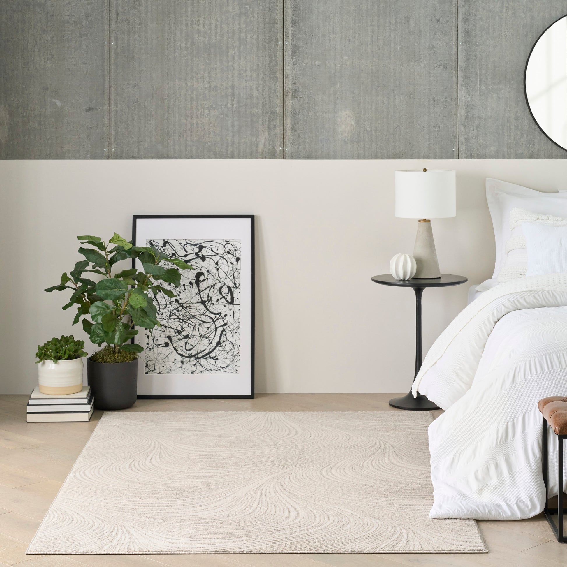 Neutral bedroom with beige woven rug, white bedding, black round side table, green potted plant, and framed abstract art.
