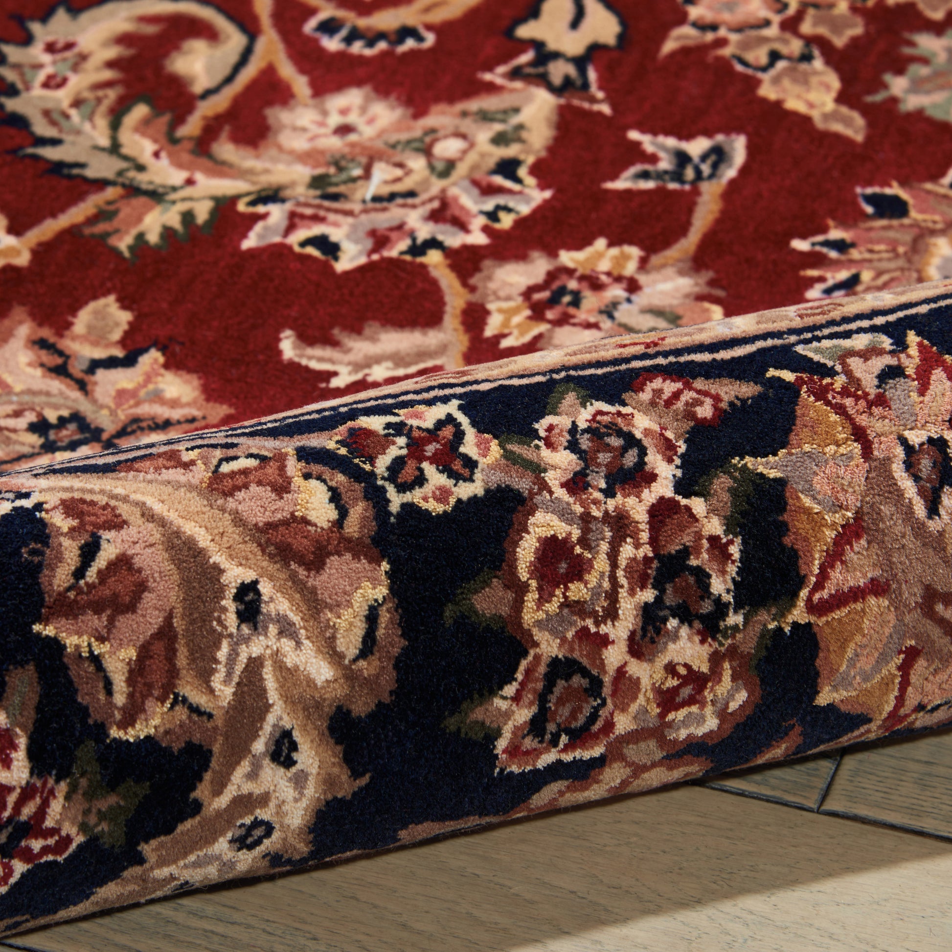 Close-up of a red oriental rug with a navy border and cream floral motifs, showing a folded edge on a wooden floor.