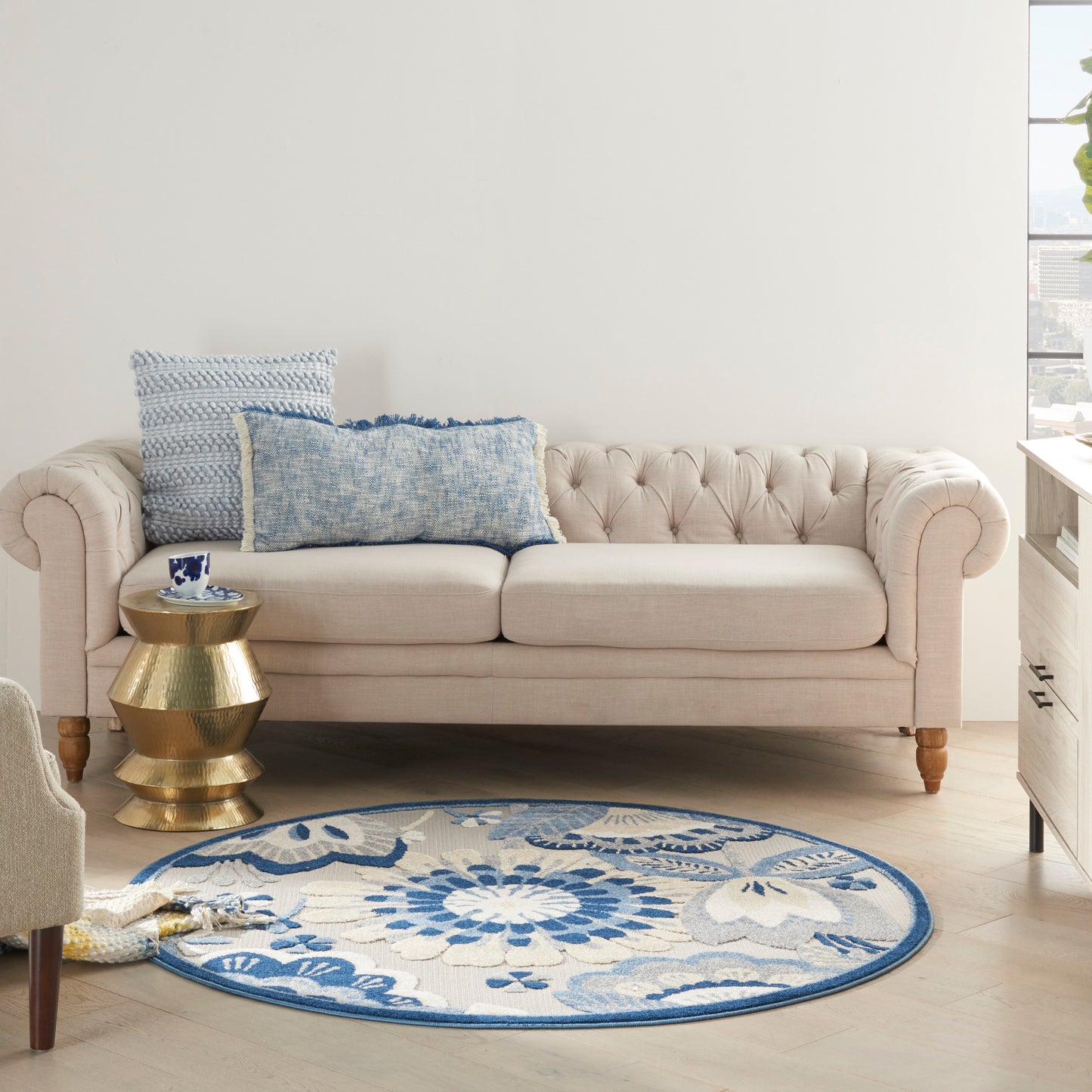 Round blue floral rug, 5' diameter, in a cozy living room with a beige tufted sofa, blue pillows, brass side table and light wood floor.