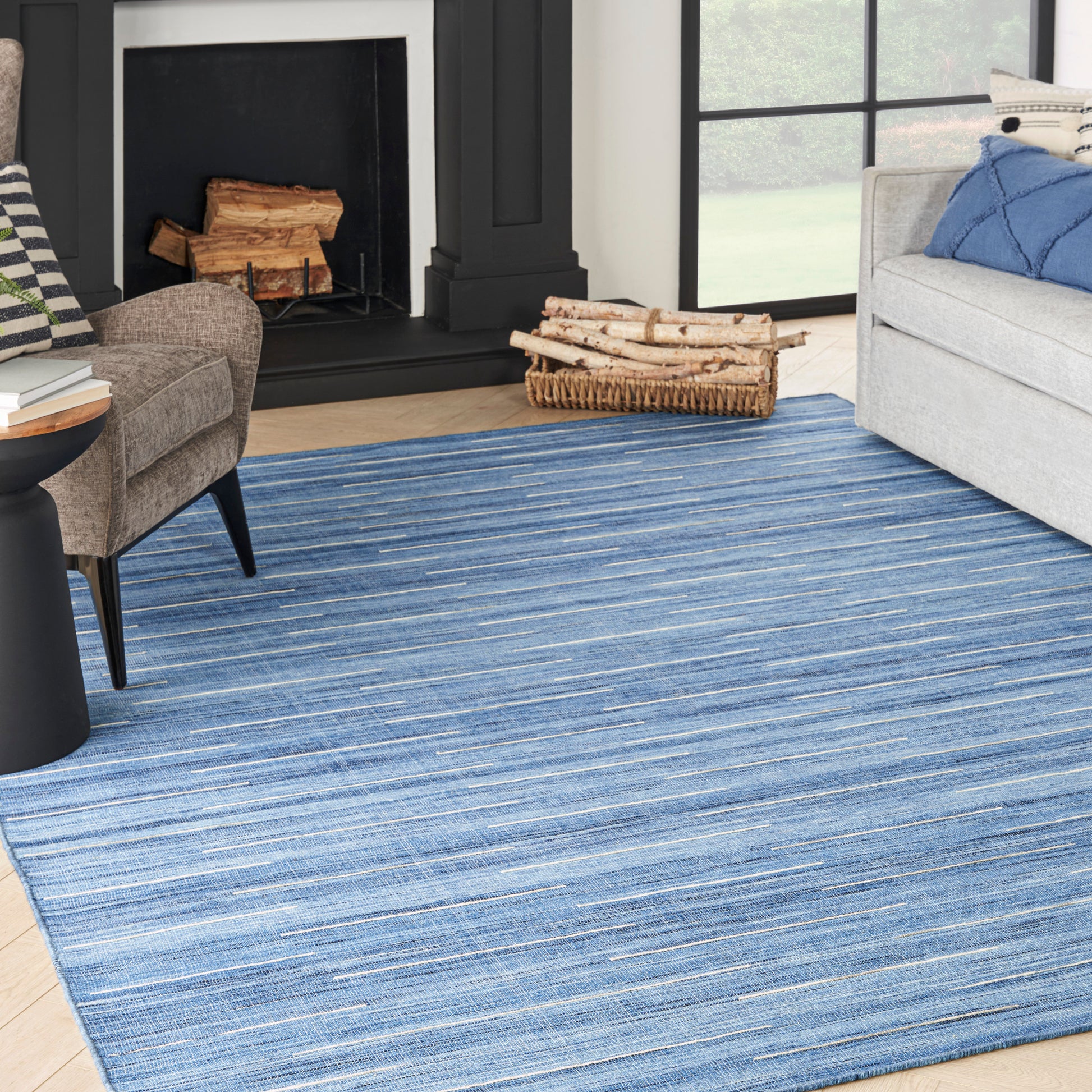 Blue striped woven area rug in a living room with a black fireplace, gray sofa, brown chair, and firewood basket.