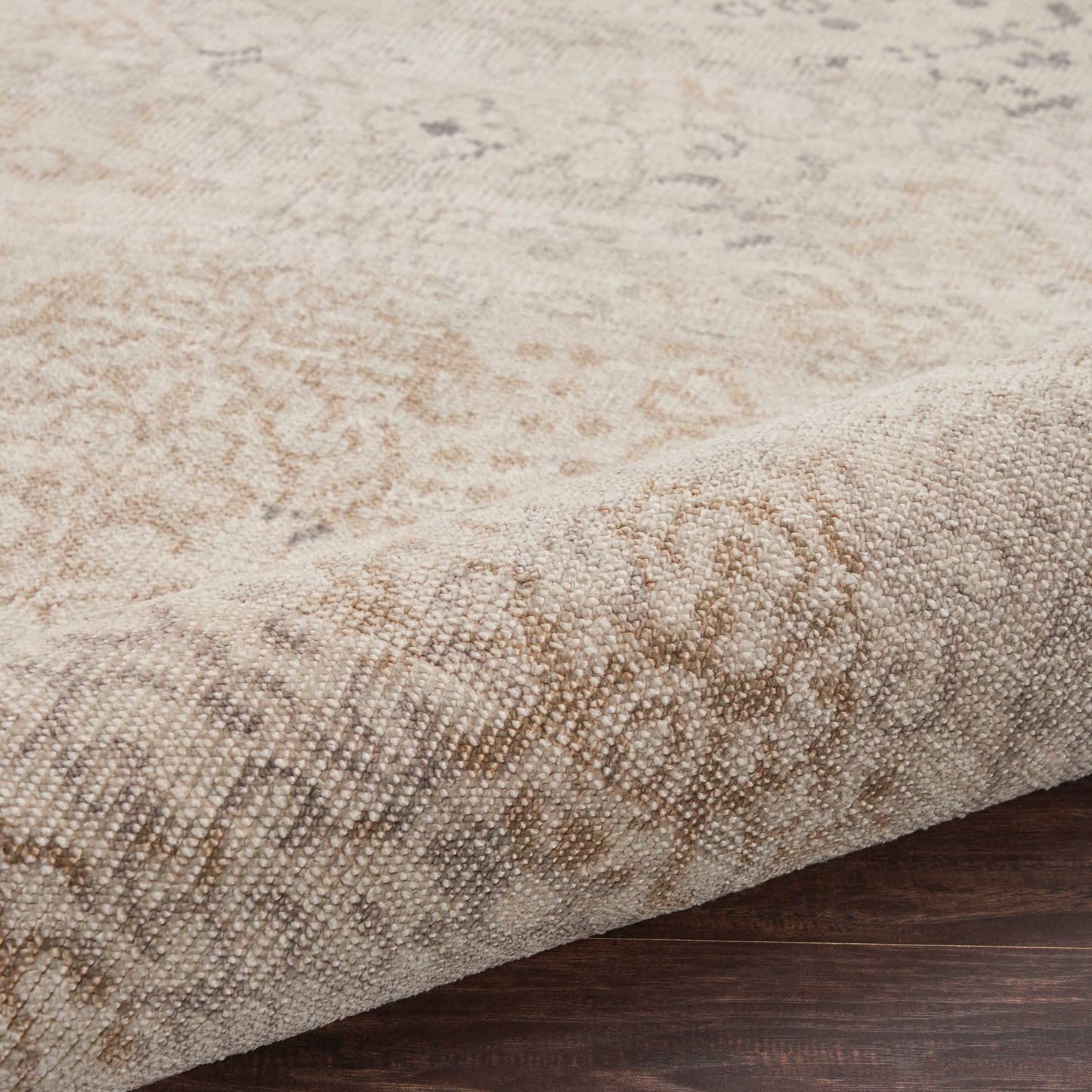 Beige woven area rug with subtle oriental medallion pattern, cream and tan tones, folded edge on dark wood floor.
