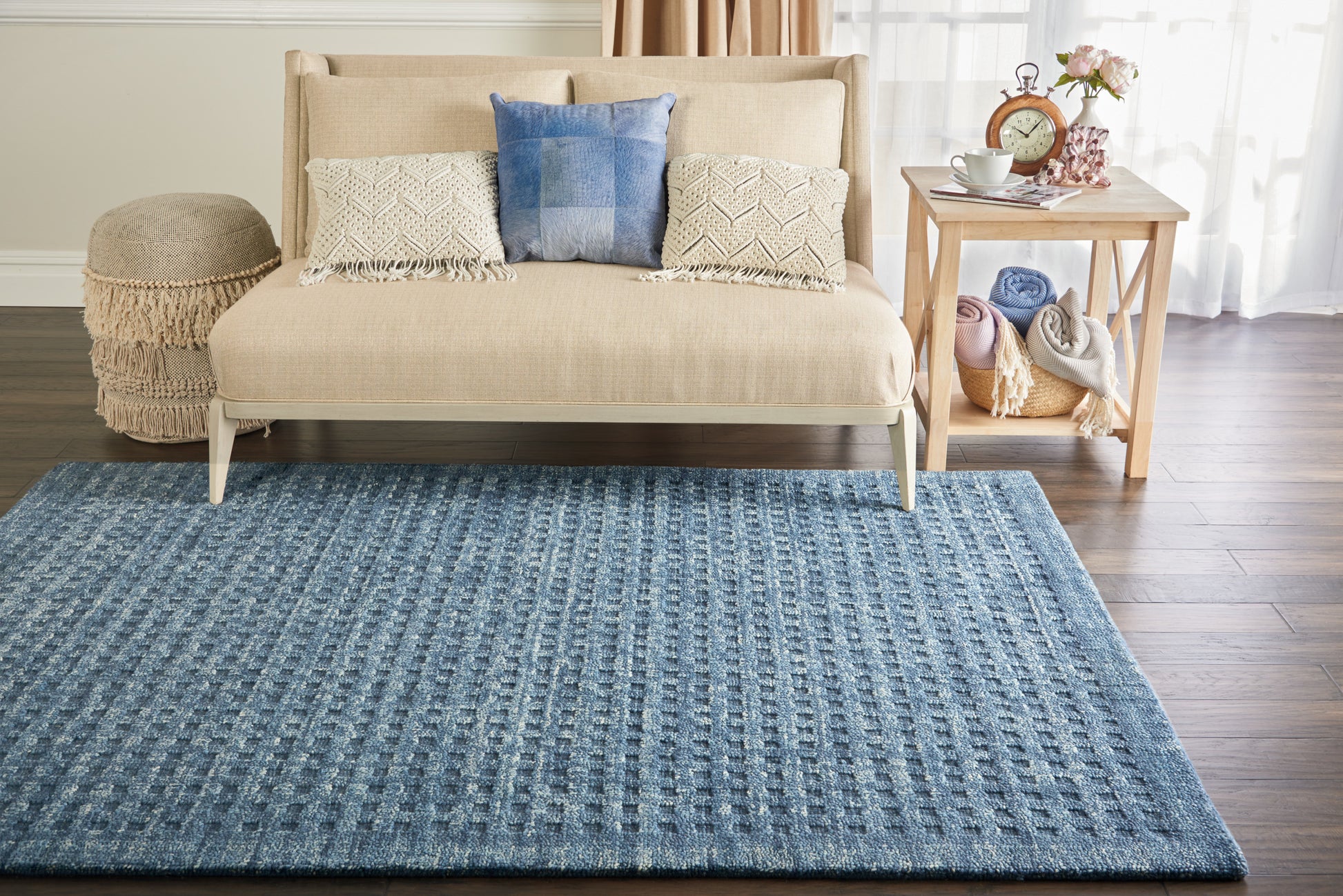 Beige sofa with blue and cream textured cushions on a blue woven rug; oak side table with blankets and flowers.