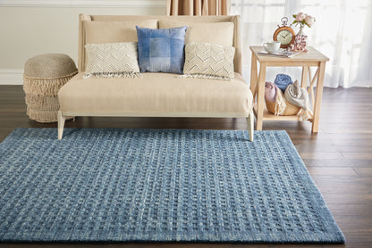 Beige sofa with blue and cream textured cushions on a blue woven rug; oak side table with blankets and flowers.