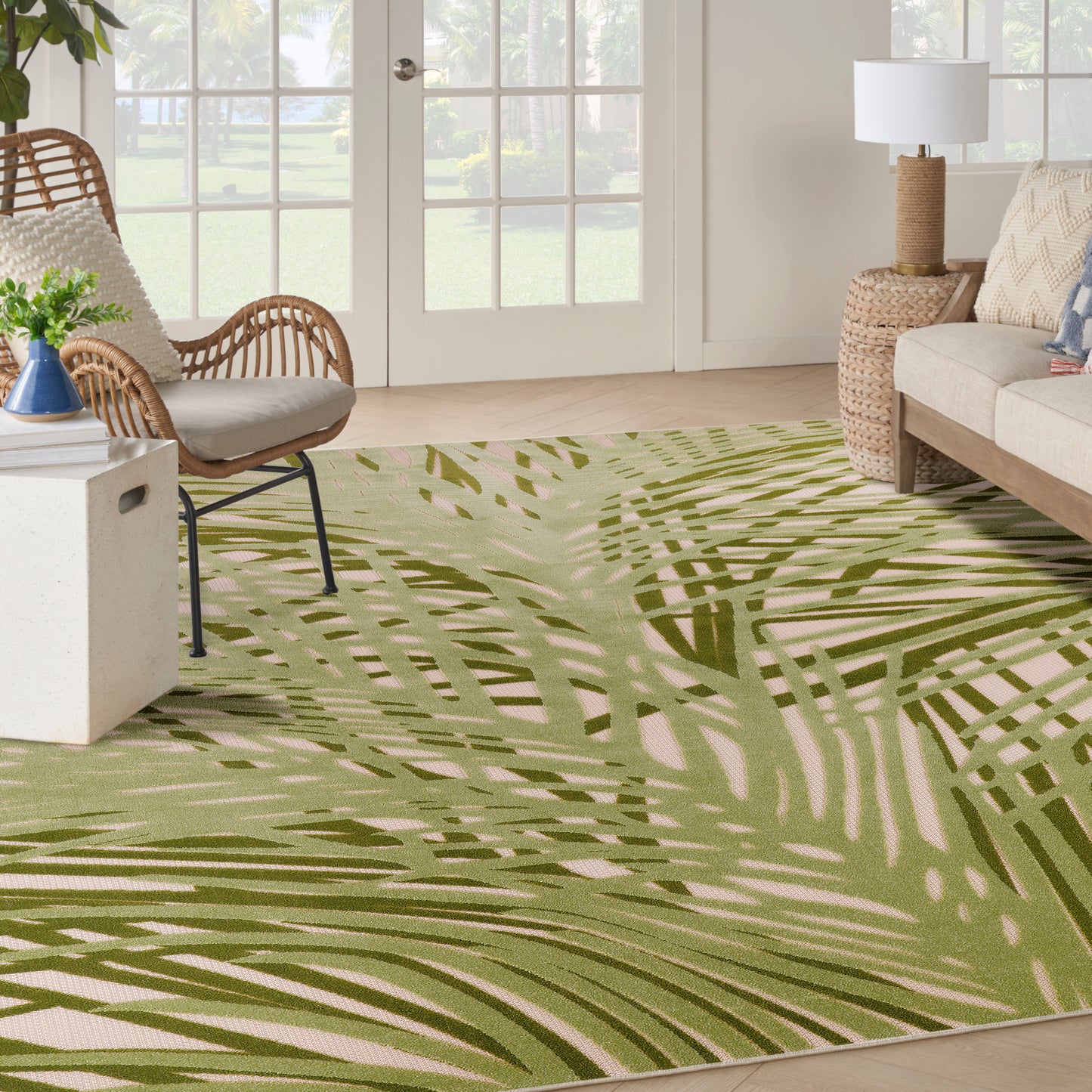 Olive-green palm-leaf patterned rug in a bright sunlit living room with wicker chairs, neutral sofa & woven side table.