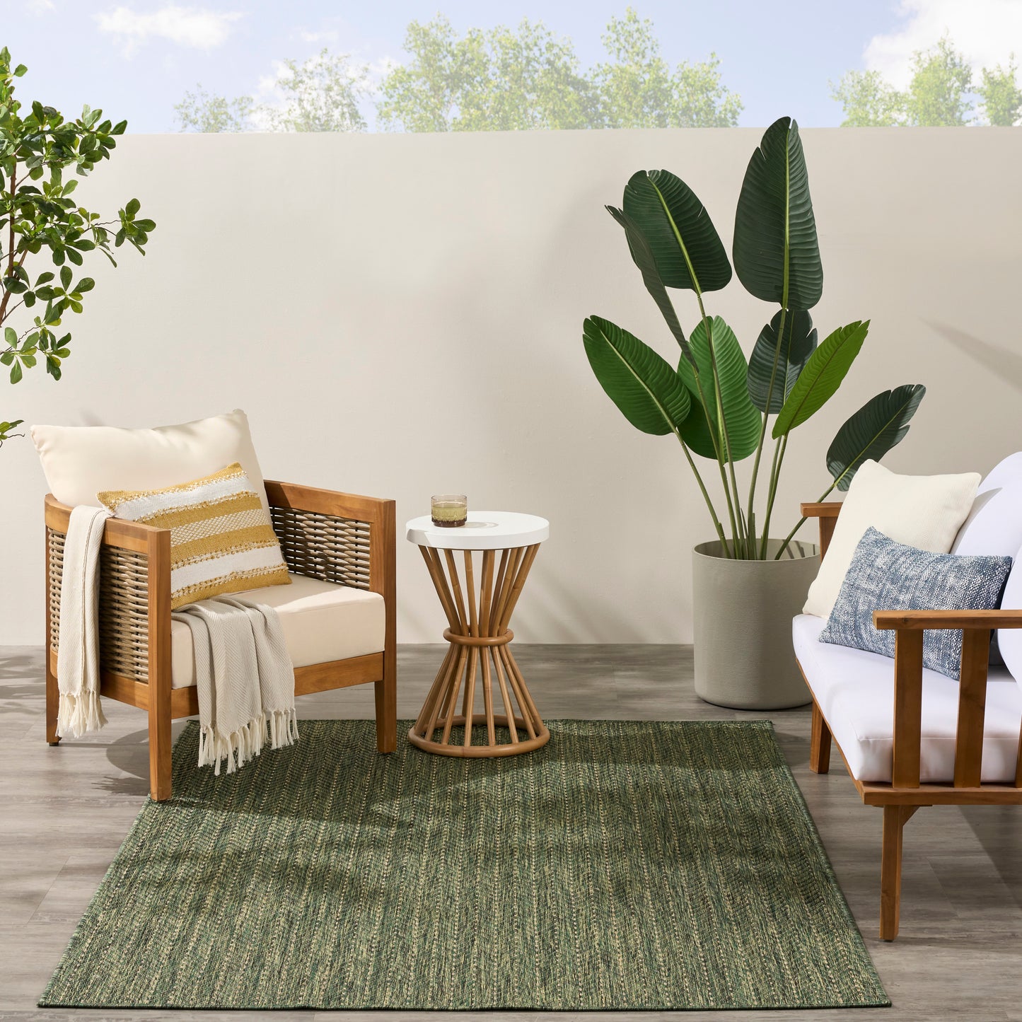Outdoor patio with wooden armchairs, white cushions, striped throw, round side table, large potted plant, and green rug.