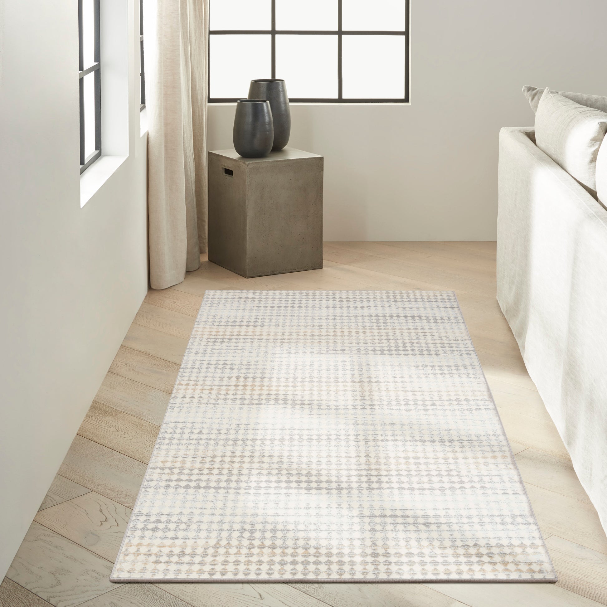 Light gray geometric rug on pale wood floor beside a fabric sofa; concrete side table with two dark vases by a window.