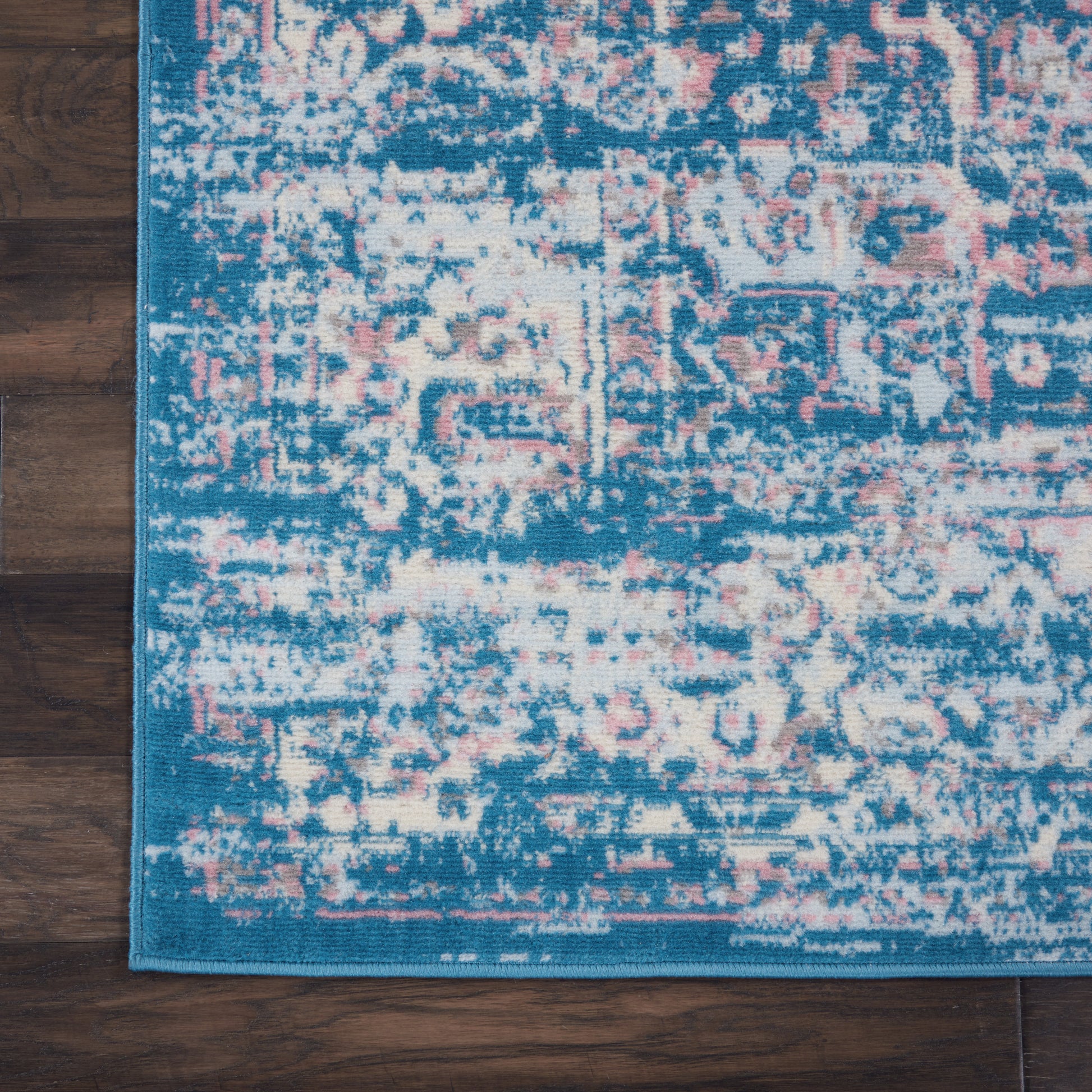 Rectangular blue distressed area rug with pink and ivory pattern on wood floor, blue border along edge.