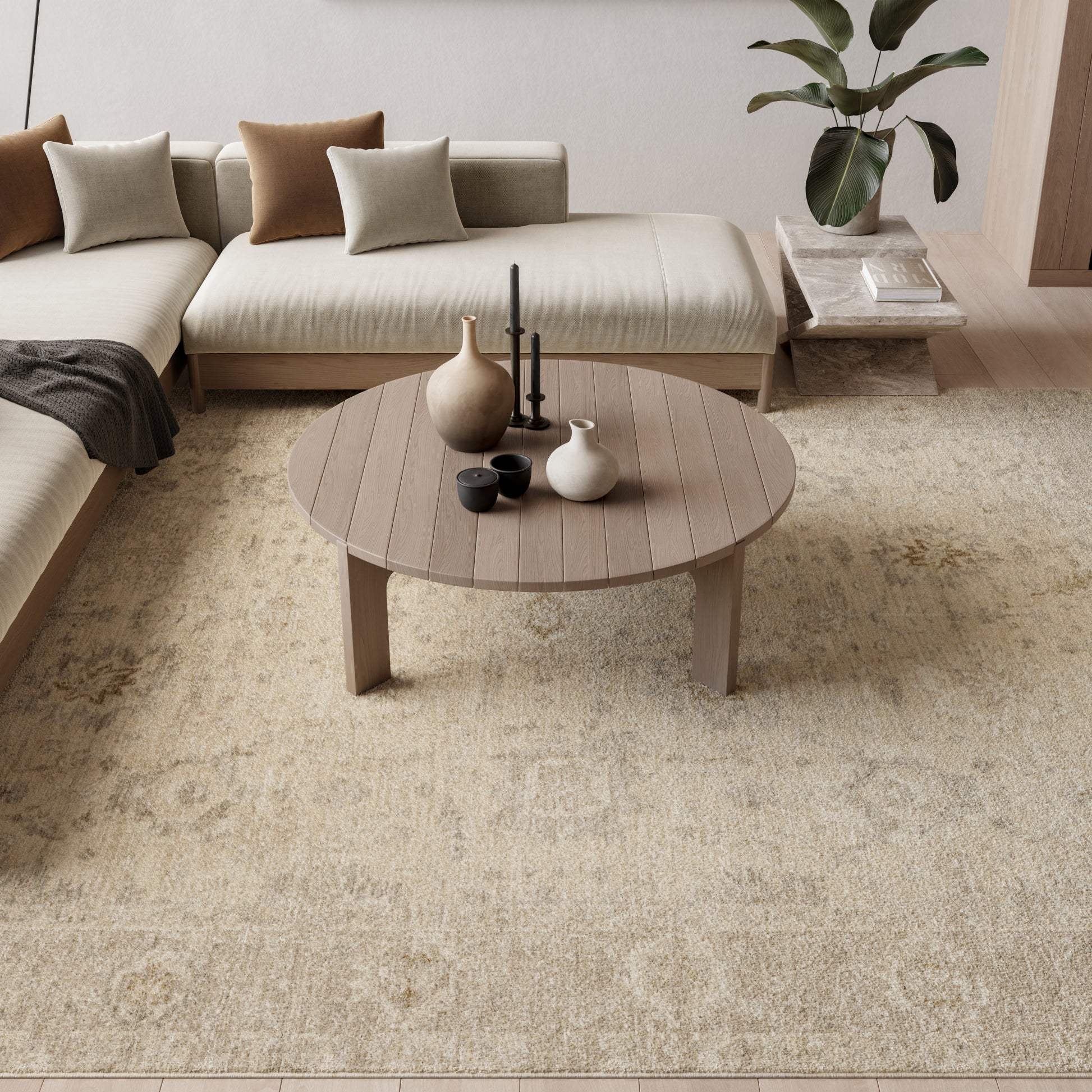 Round wooden coffee table on a beige rug in a neutral living room with a sectional sofa, cushions and a potted plant.