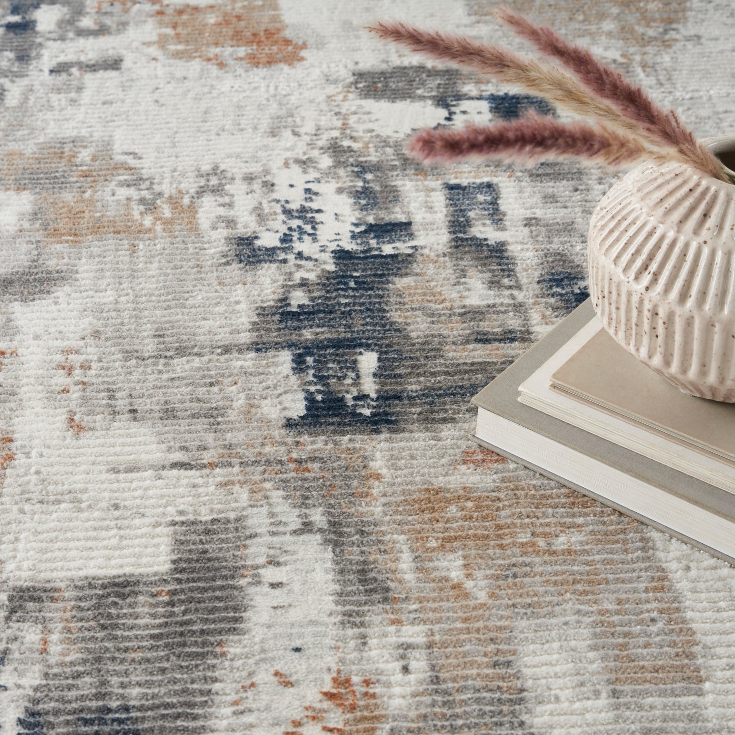 Blue-beige abstract rug in neutral tones, with a cream ribbed ceramic vase containing pampas grass atop neutral stacked books.