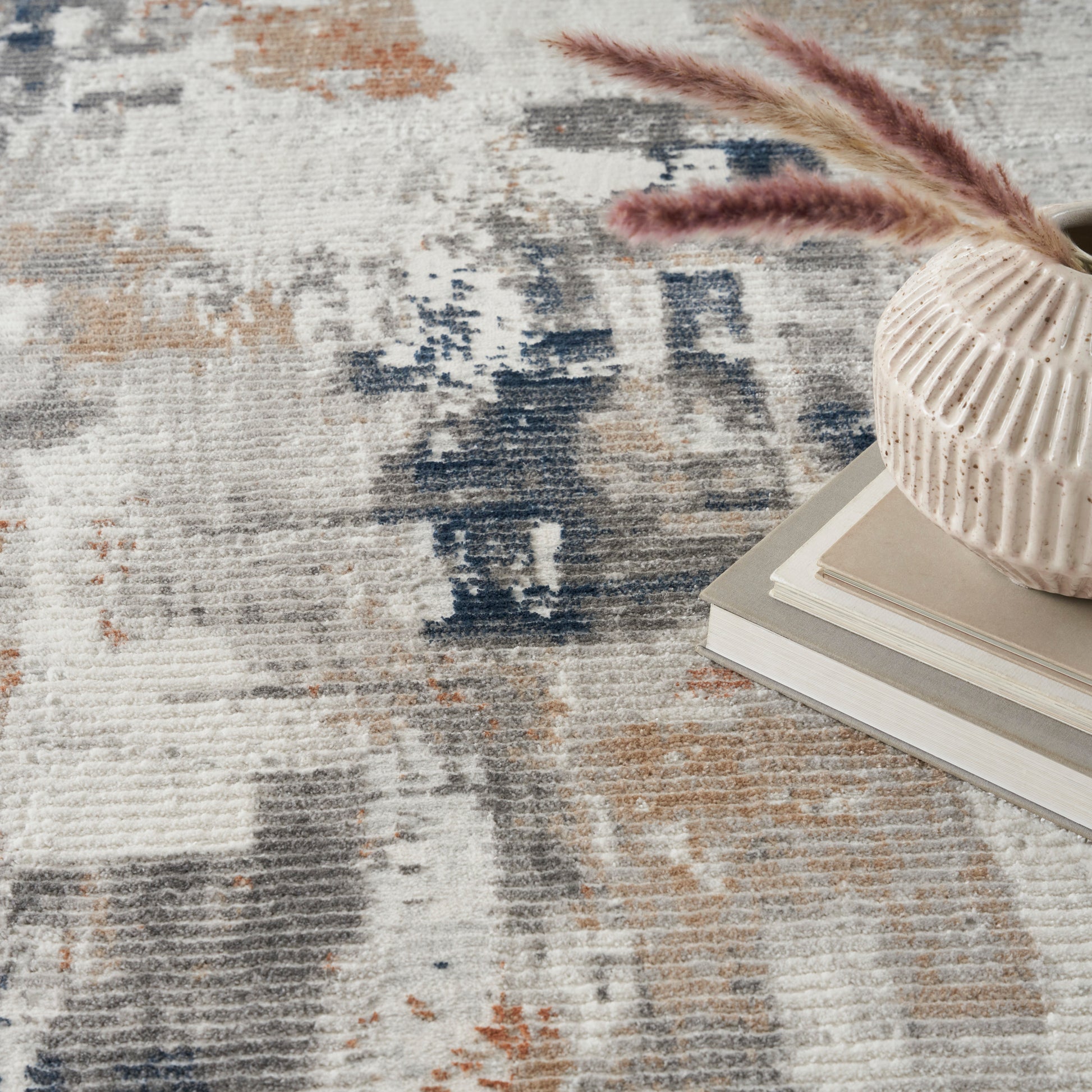 Blue-beige abstract rug in neutral tones, with a cream ribbed ceramic vase containing pampas grass atop neutral stacked books.
