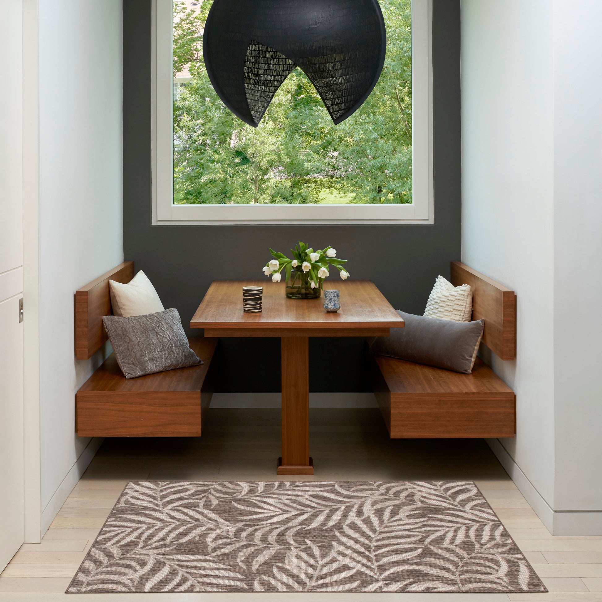 Walnut corner dining nook & square pedestal table, leaf-pattern rug, white cushions, vase of white flowers, dark wall.
