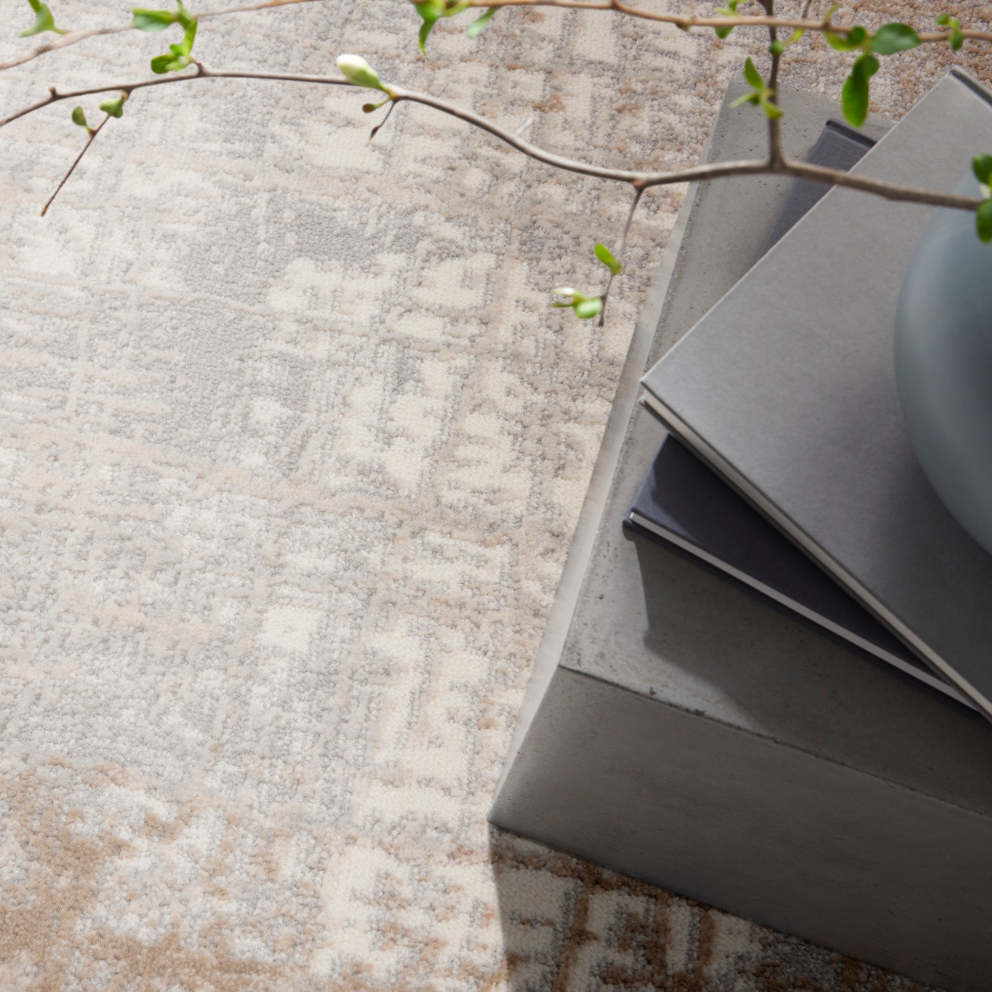 Beige abstract rug; stacked gray books on a concrete pedestal, round gray vase, and a green leafy branch.