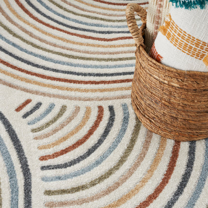 Round multicolor rug with curved concentric arc patterns in blues, oranges, beiges; wicker basket with striped throw.