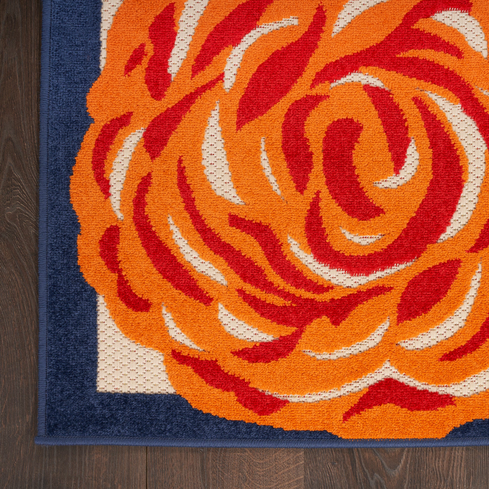 Close-up of a decorative area rug: bright orange-red rose pattern on cream with a navy border, textured loop pile on wood floor.