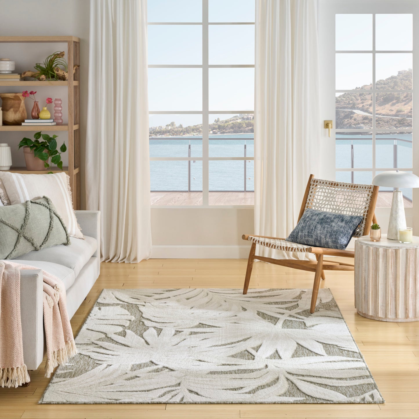 Bright coastal living room with leaf-pattern rug, light wood floors, white sofa, woven chair, plants and sea-view windows.