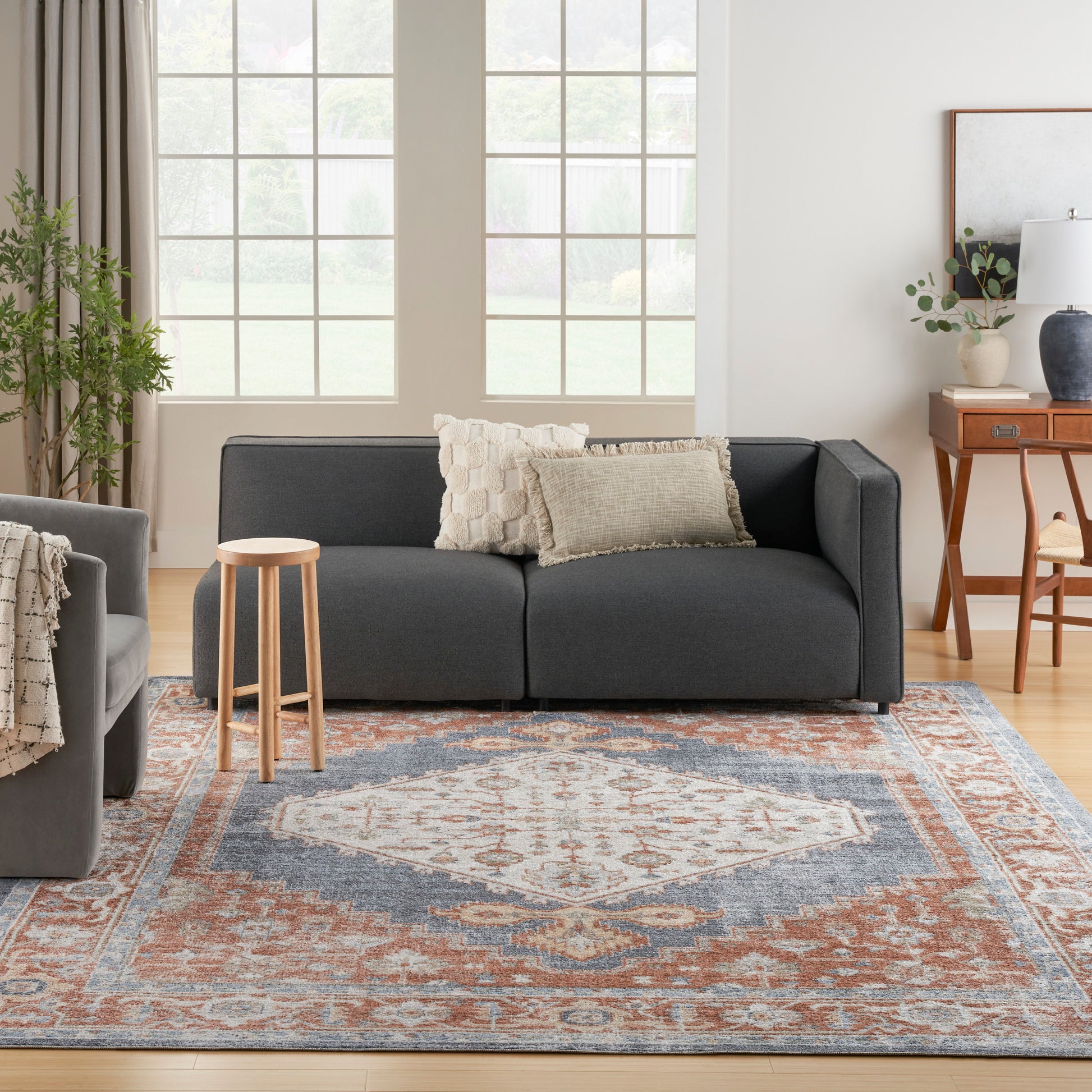 Blue-rust patterned rug in a bright living room with a dark gray modular sofa, textured beige cushions, and a light wood stool.
