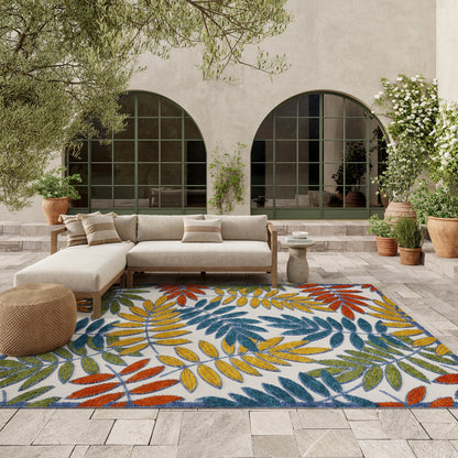Outdoor courtyard with a leaf-pattern rug in blue, yellow, red; beige sectional sofa, potted plants, arched doors.