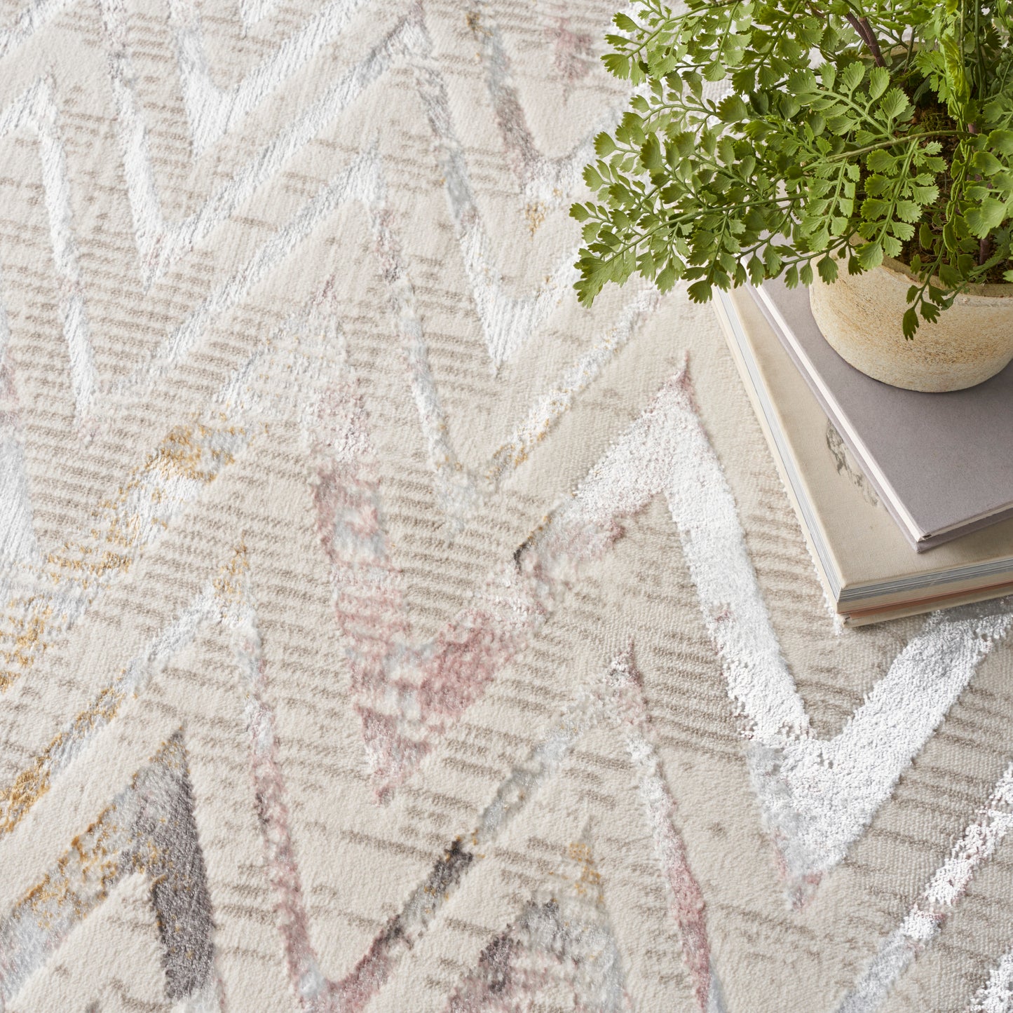 Beige woven area rug with light chevron zigzags in cream, pink and gray; potted fern atop a stack of books.