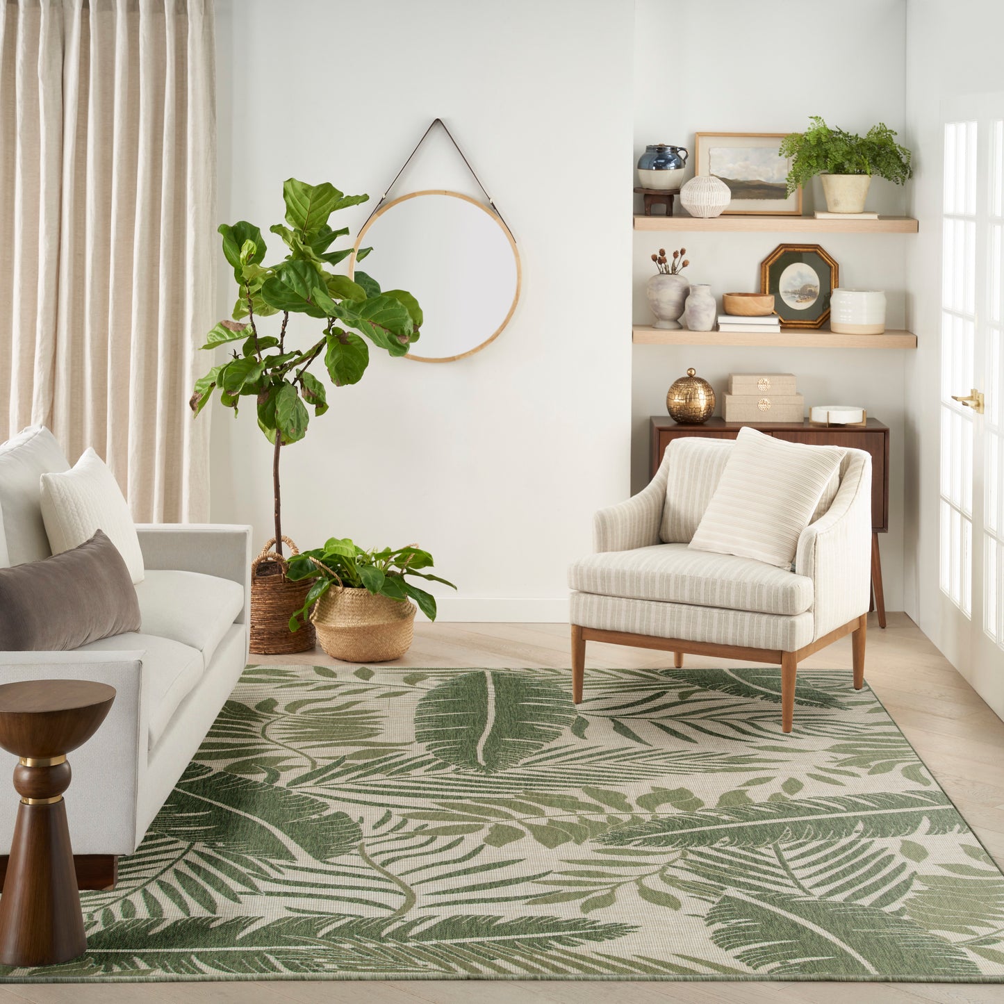 Bright living room with green palm-leaf rug, cream sofa and striped armchair, round mirror, tall plant in basket, shelves.