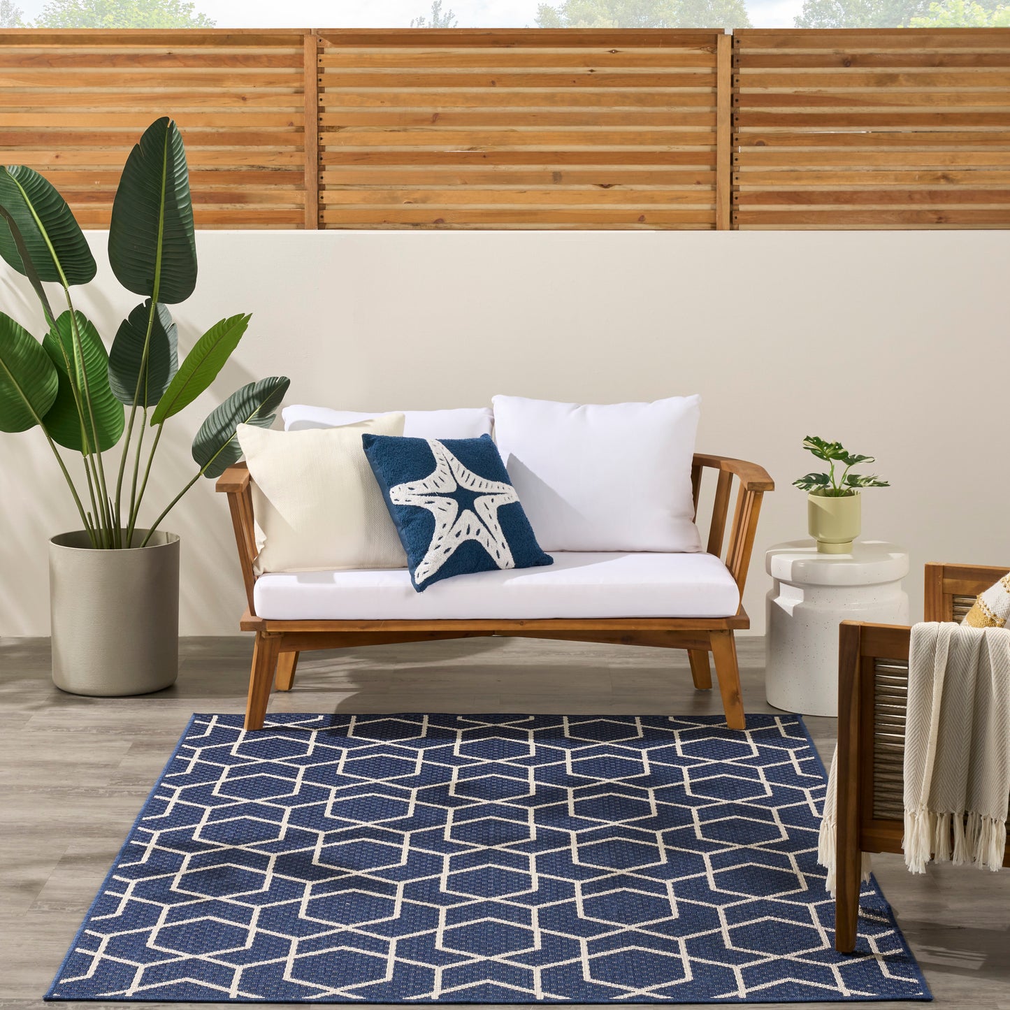 5' x 7' modern outdoor rug with a navy geometric pattern in a seating area, featuring a wood-framed sofa with white cushions, navy starfish pillow, beige pillow, and a potted plant.