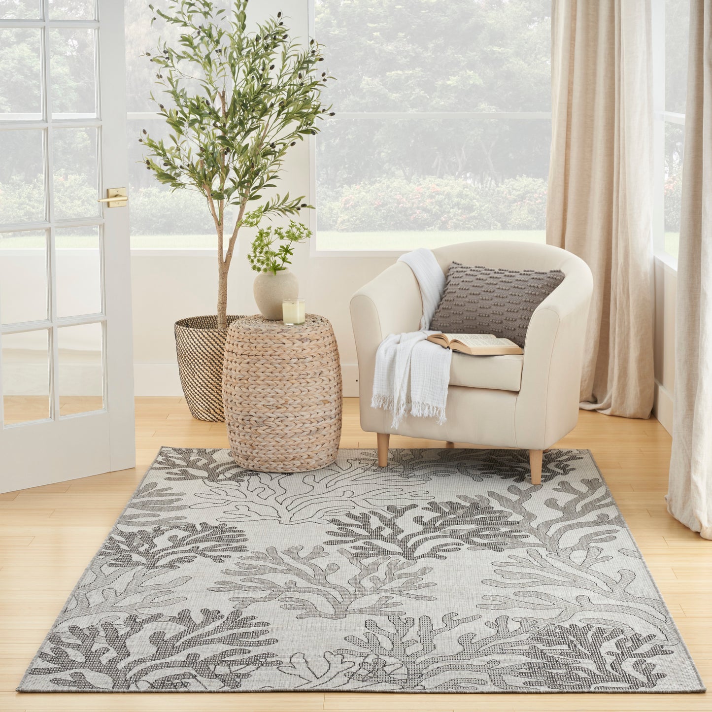 Beige upholstered armchair beside woven jute tables, gray botanical rug, potted plants, candle, cozy living corner.
