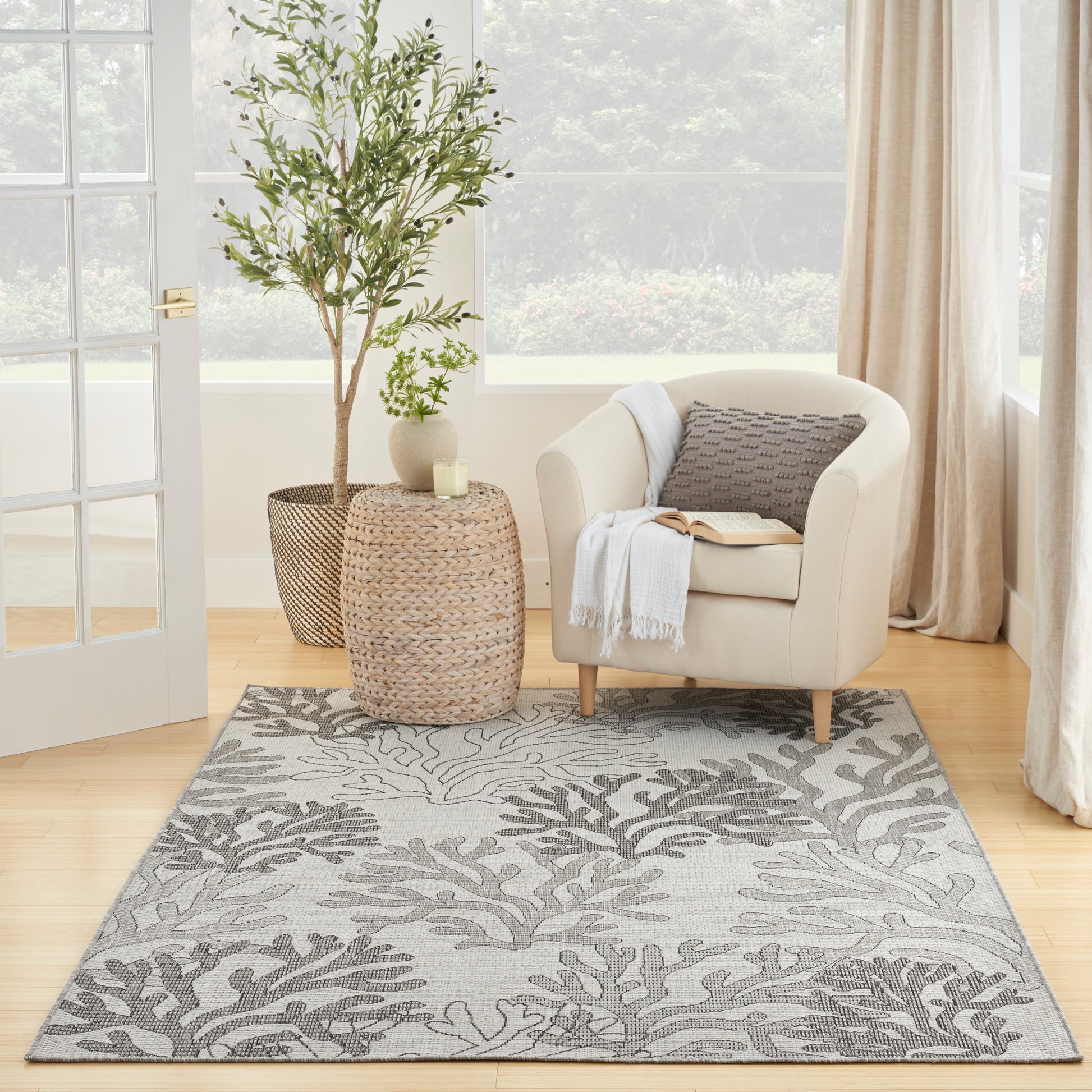 Beige upholstered armchair beside woven jute tables, gray botanical rug, potted plants, candle, cozy living corner.