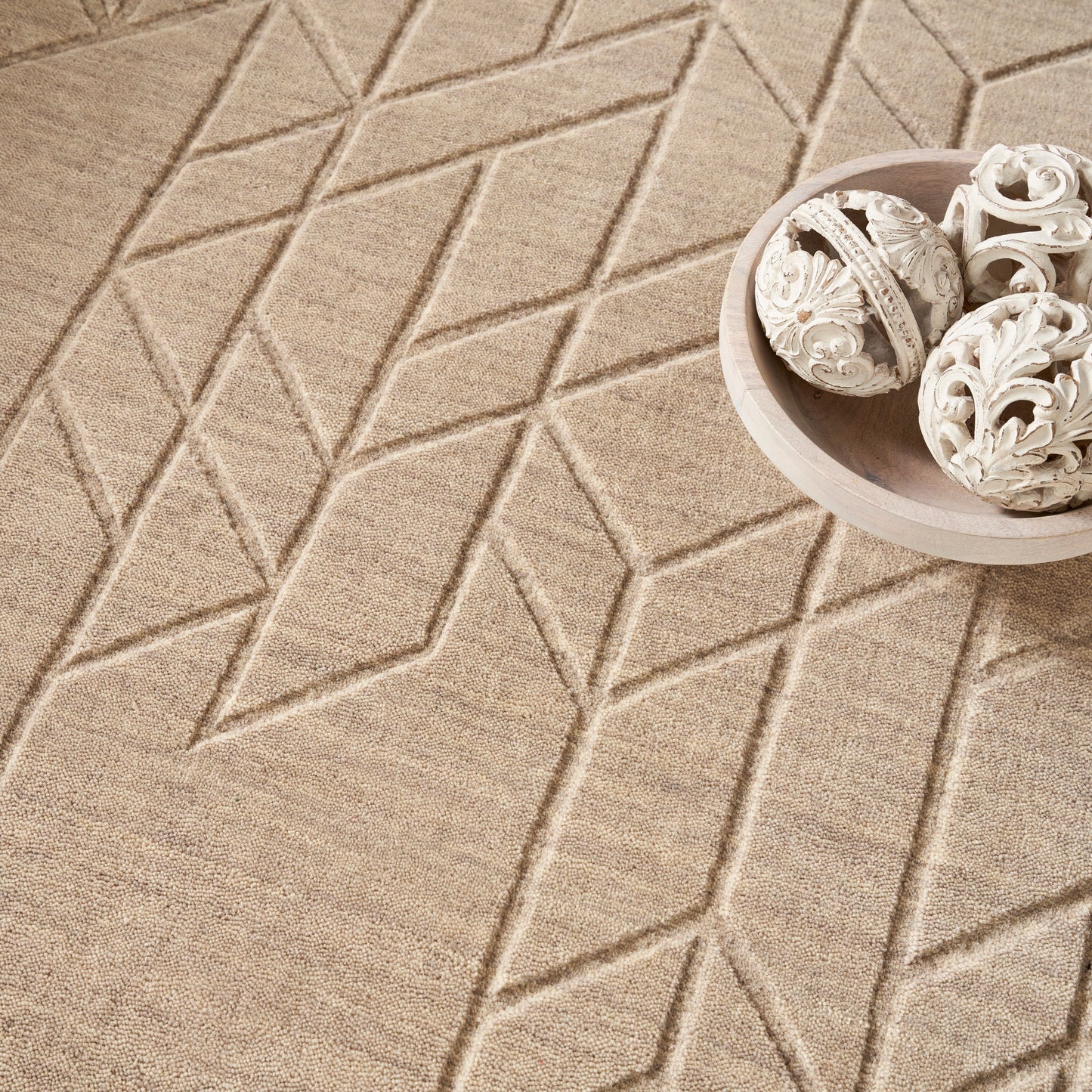 Beige geometric rug with raised diamond pattern; round light wood bowl containing white carved ornamental spheres.