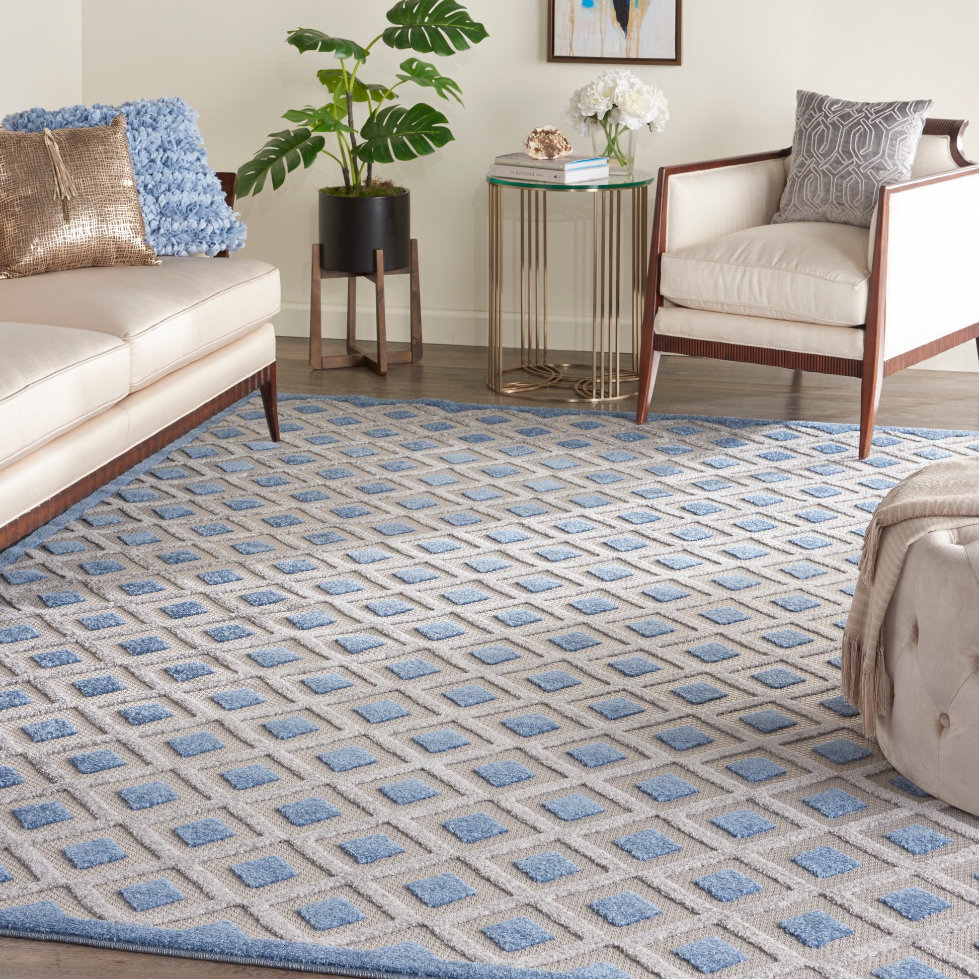Living room with a blue and beige diamond-pattern rug, cream sofa, gold-accent cushions, wooden chairs, plant and glass-top side table.