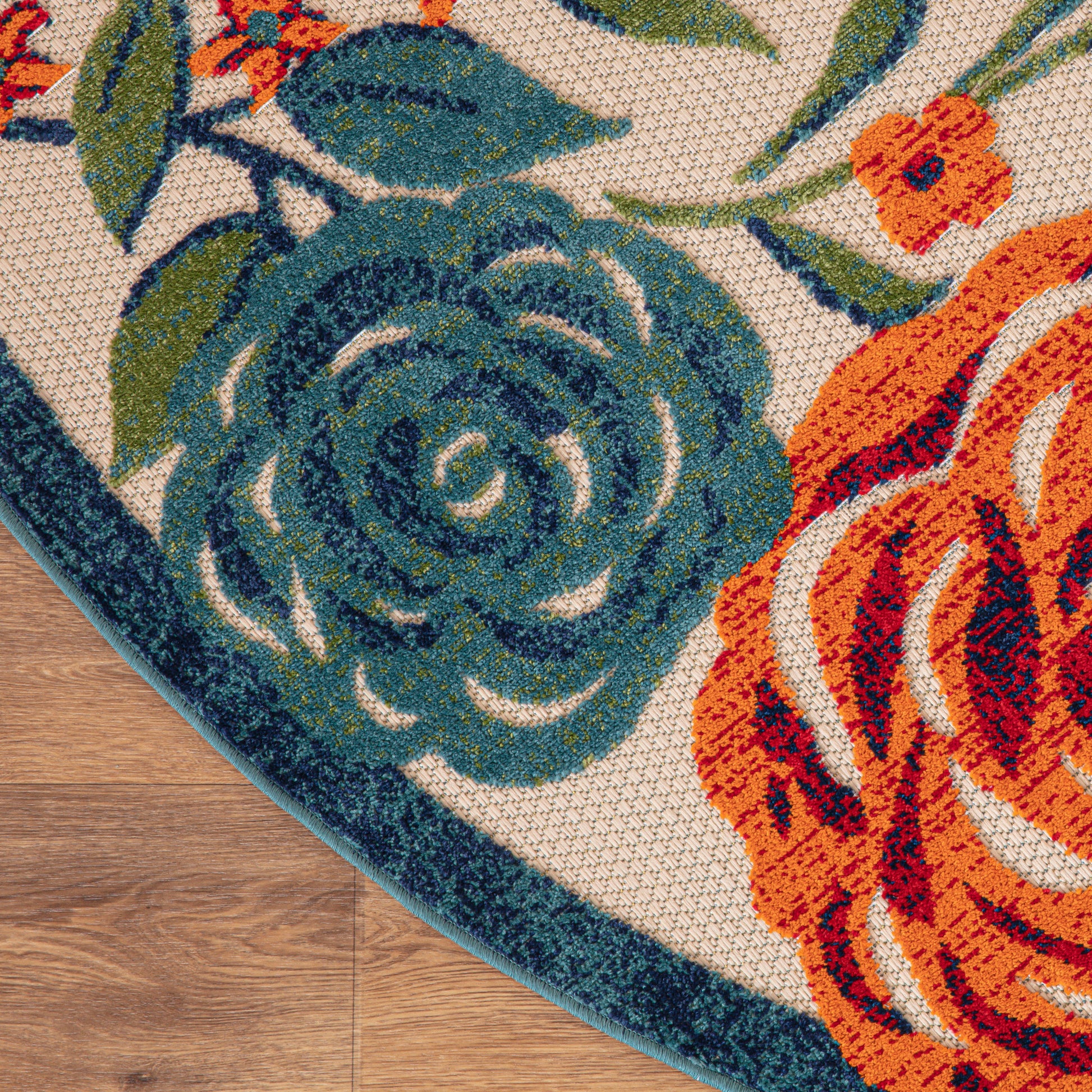 Round outdoor rug with teal blue rose and orange floral motifs on beige weave, teal edge, laid on wood floor.