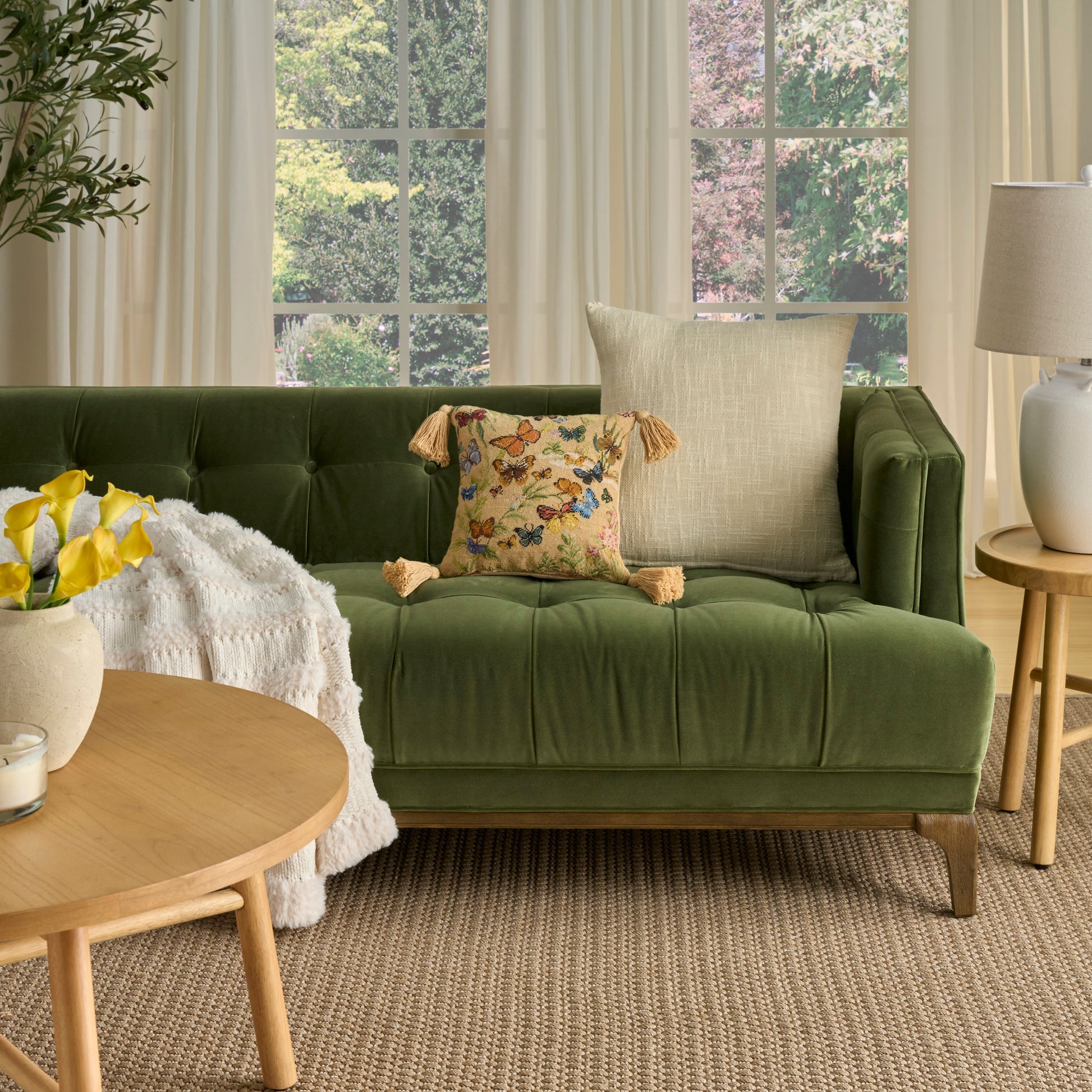 Olive green velvet tufted sofa with tassel pillow; round wood coffee table, yellow flowers, cream throw, natural rug.