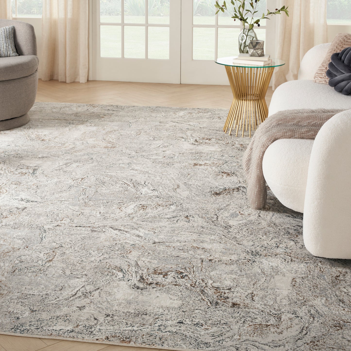 Living room with abstract gray-beige rug, cream curved sofa, gray accent chair, and glass-top gold side table by sheer curtains.
