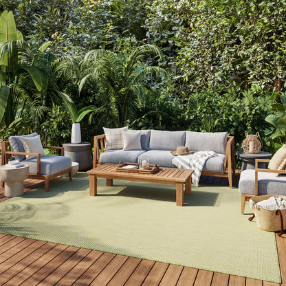 Outdoor wooden-frame sofa and chairs with gray cushions, coffee table, beige throw, on a light green rug amid lush greenery.