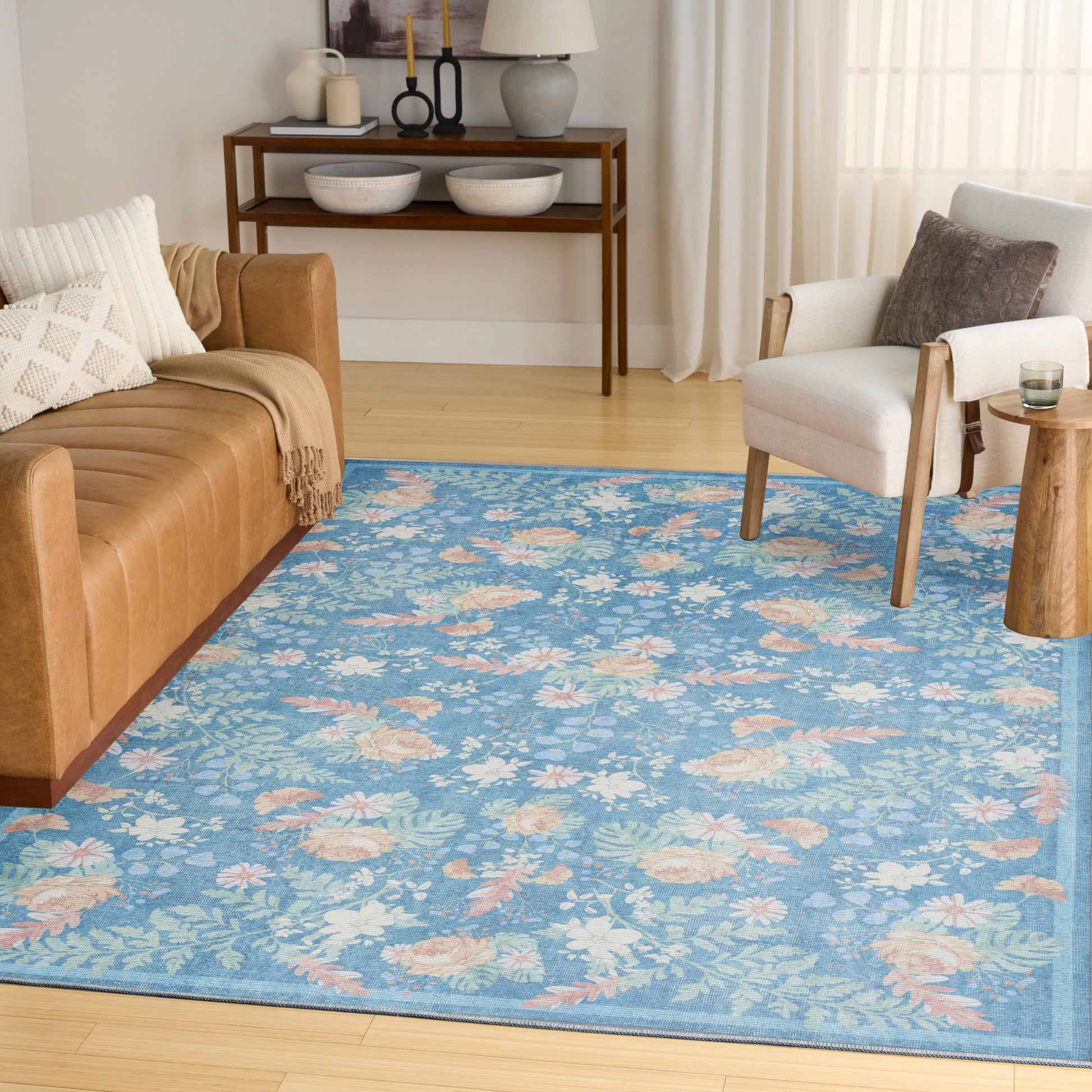 Blue floral area rug over light hardwood floor with tan leather sofa, gray accent chair, and wooden console table.