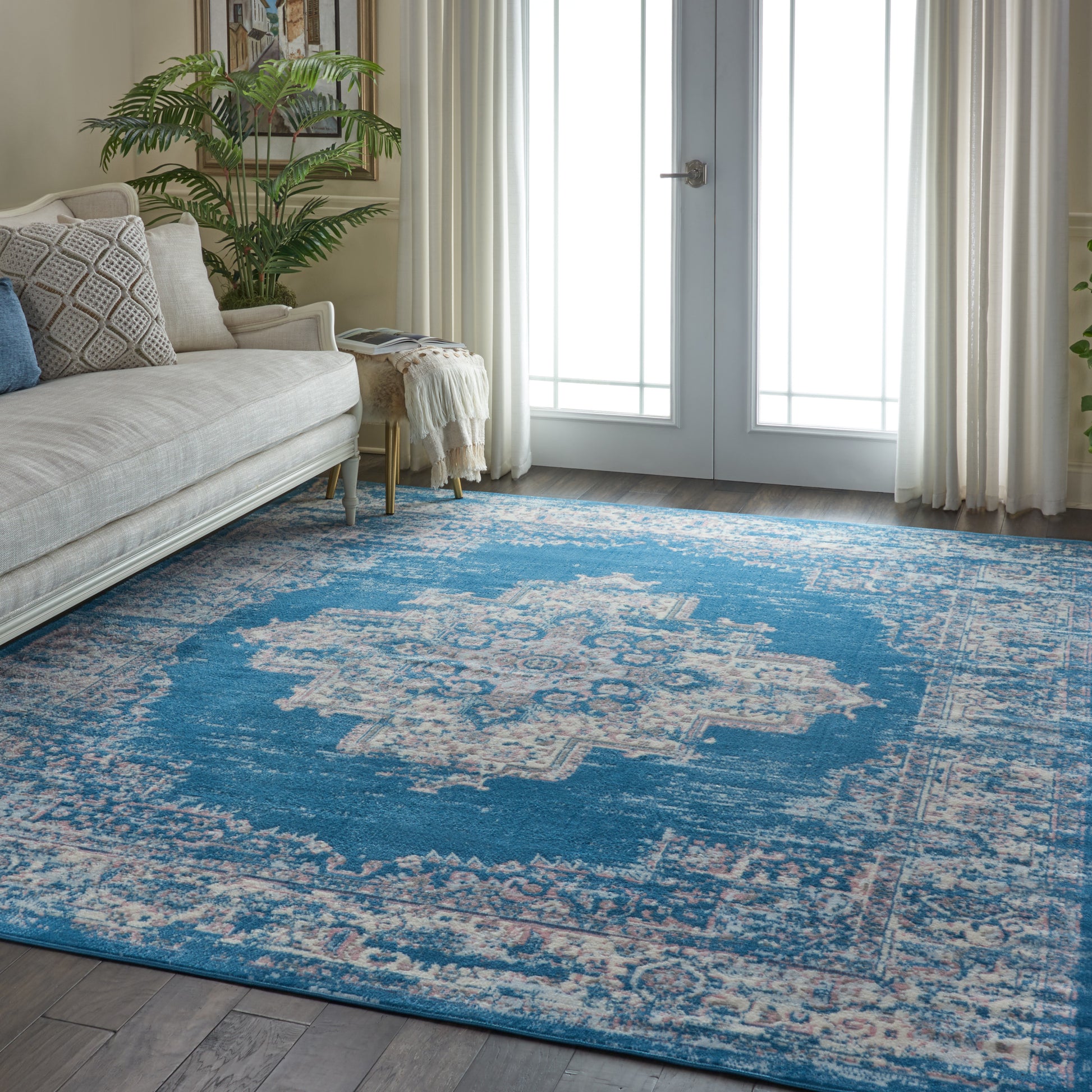 Blue patterned area rug on wooden floor beside beige sofa with cushions, potted plant, and white curtains by French doors.