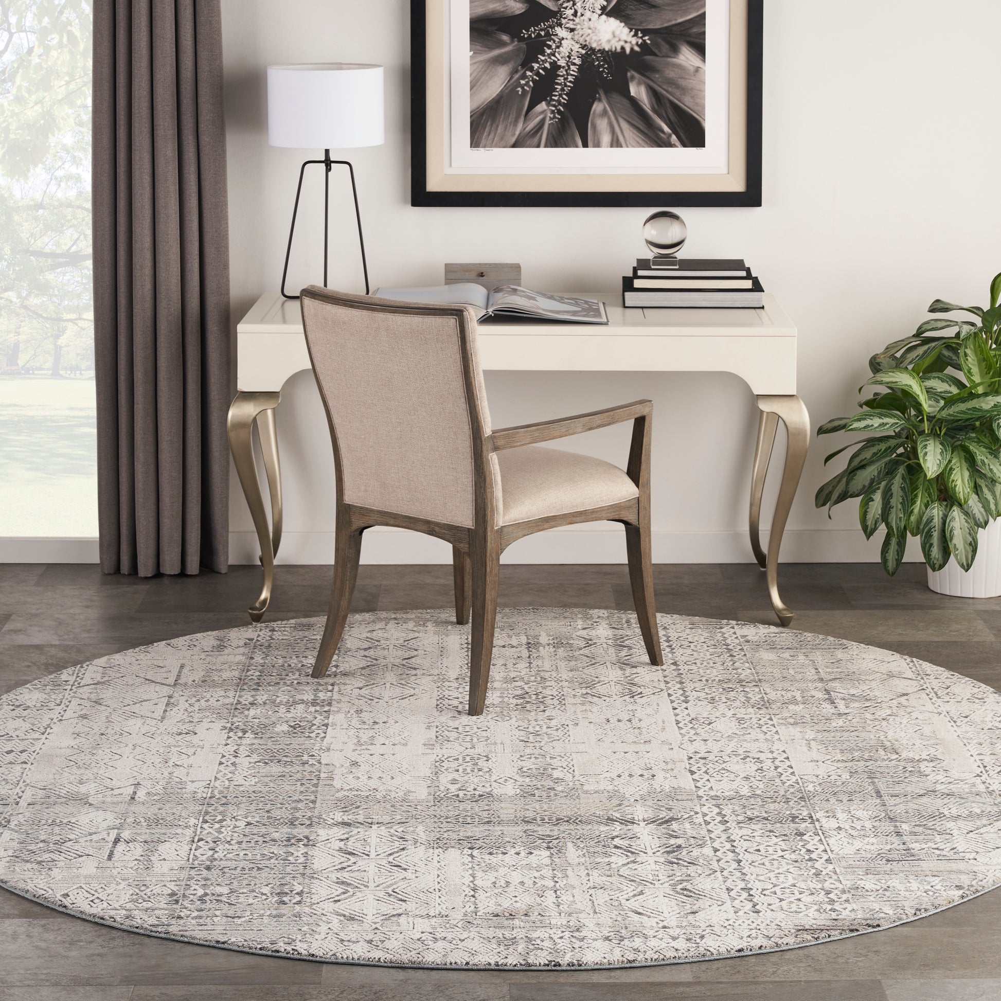 Beige upholstered desk chair with wooden frame at a white desk, round gray patterned rug, lamp, plant and framed art.