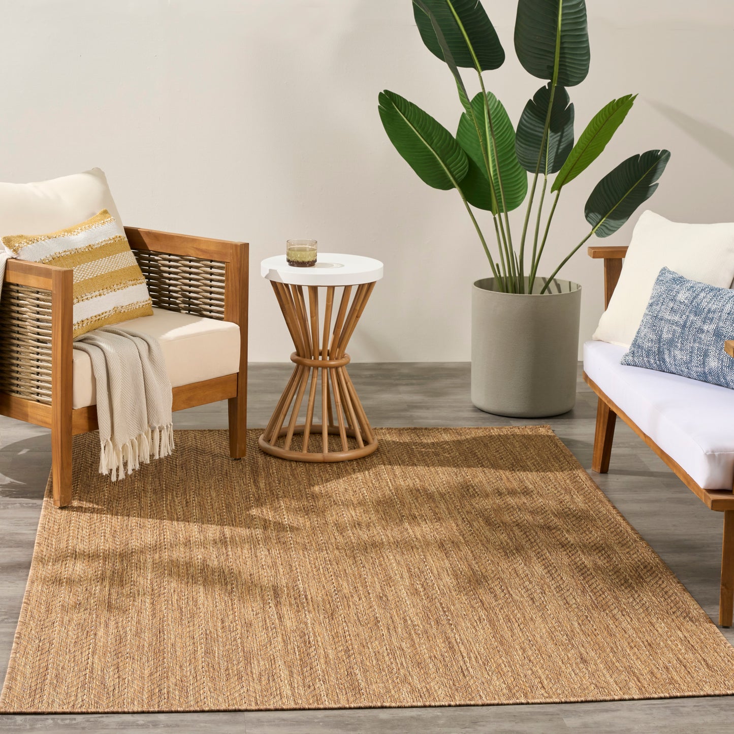 Two wood-framed armchairs with white cushions, round white-top wicker table, beige jute rug, tall potted tropical plant.