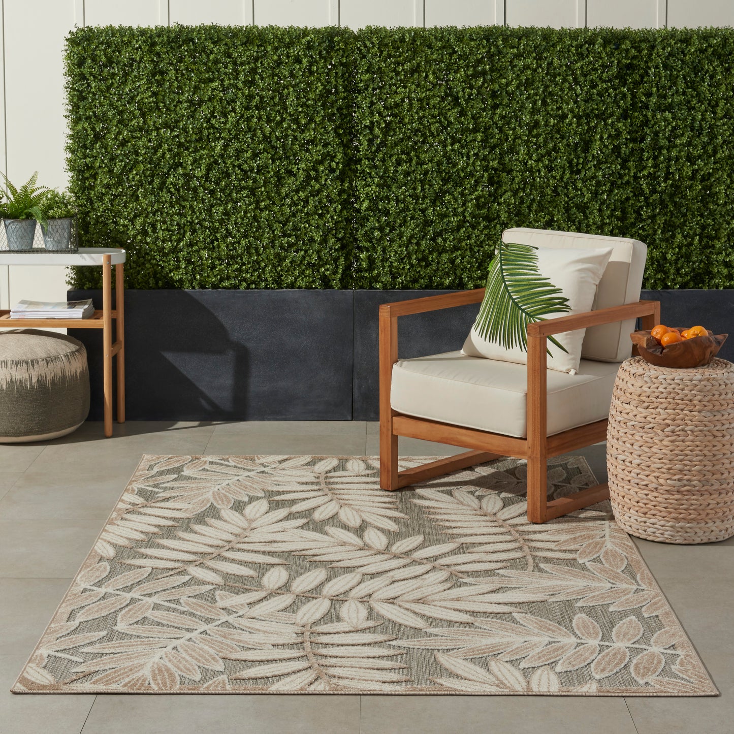 Outdoor seating area with beige cushioned wooden chair, palm leaf pillow, woven basket side table & beige leaf-pattern outdoor rug.