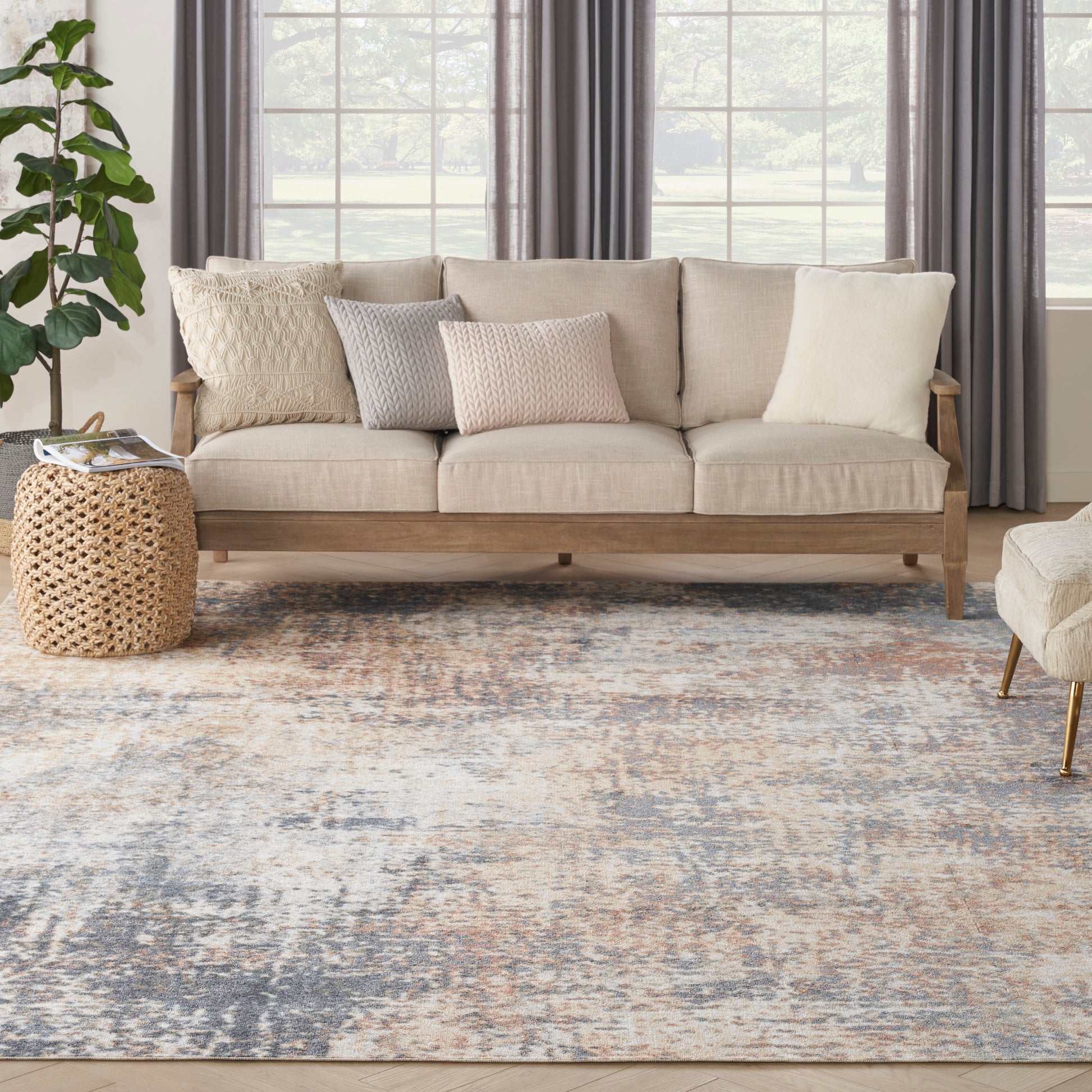 Beige fabric sofa with wooden frame, neutral knit pillows, woven rattan side table, blue-gray abstract rug, potted plant.